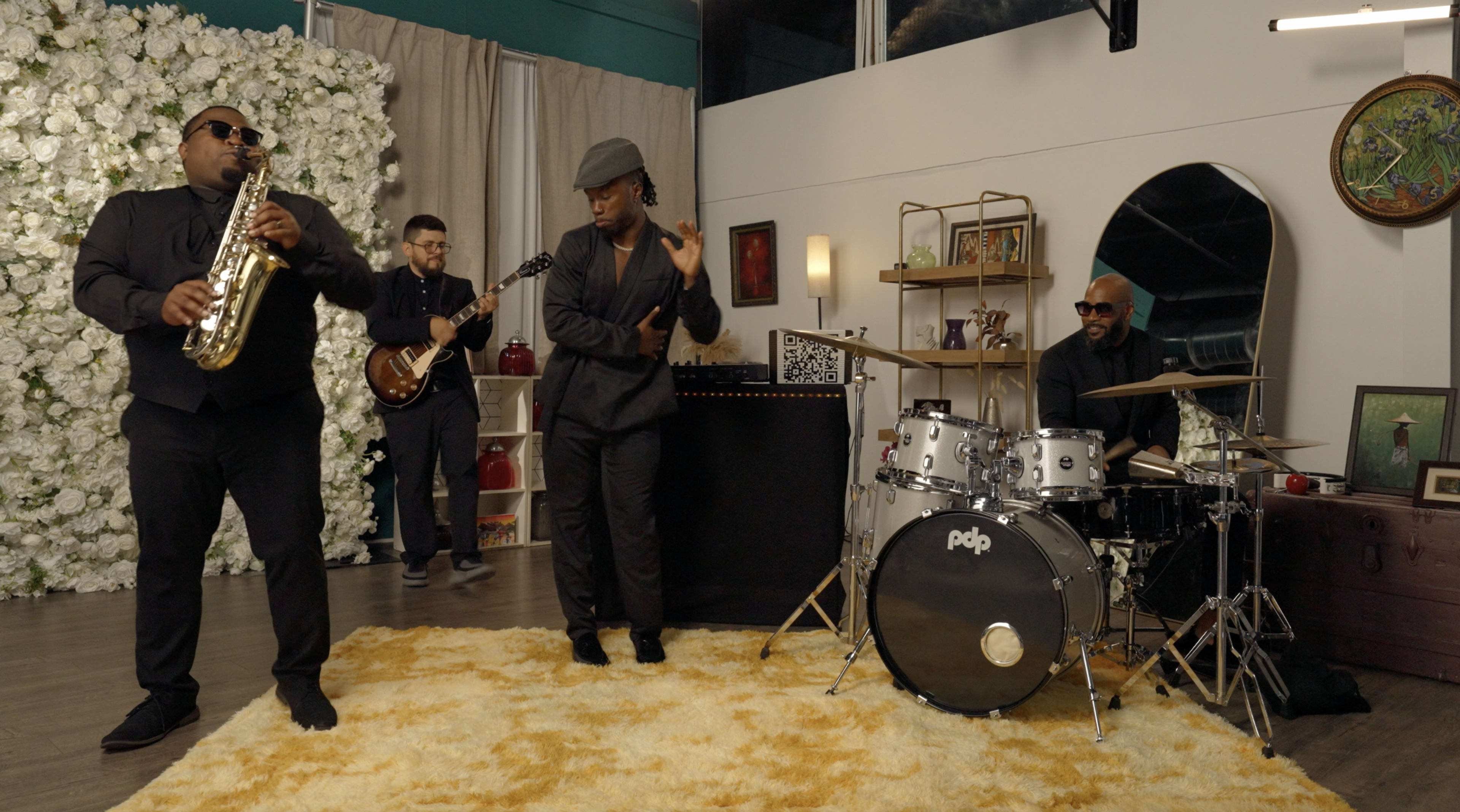 A band performs in a decorated indoor space with a floral backdrop, featuring a saxophonist, a guitarist, a percussionist, and a drummer.