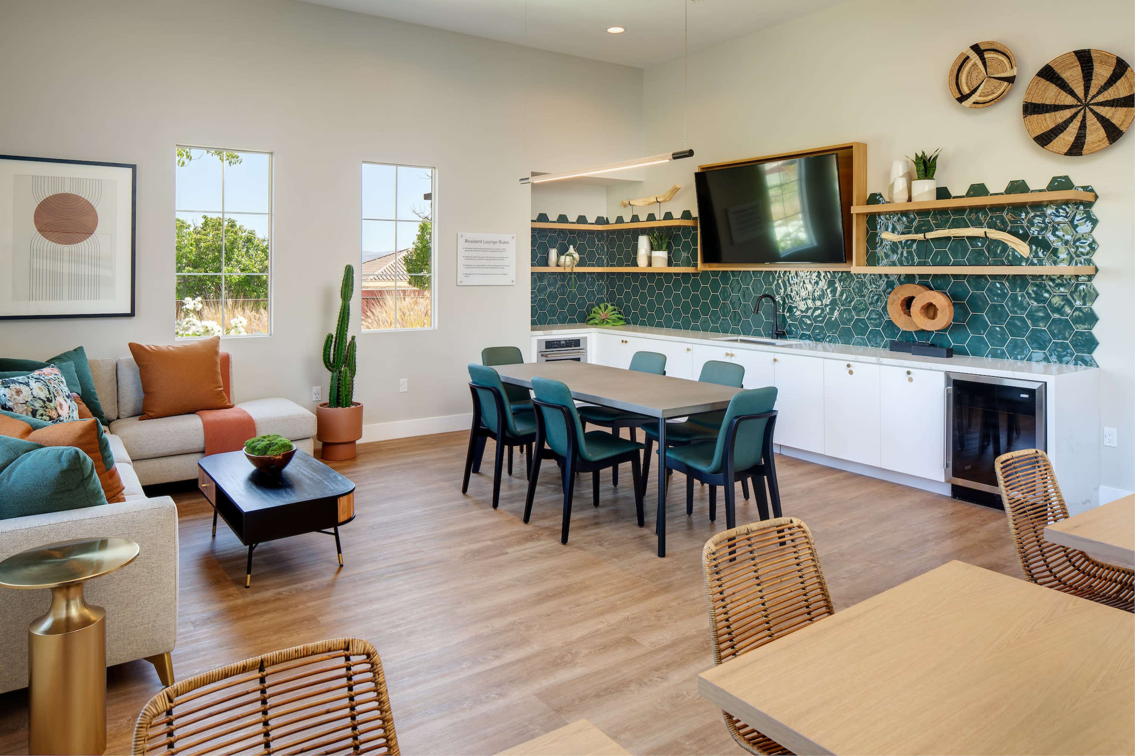 A bright, modern lounge area features a sofa, a dining table with chairs, and a kitchen area with green tile and decorative wall items.