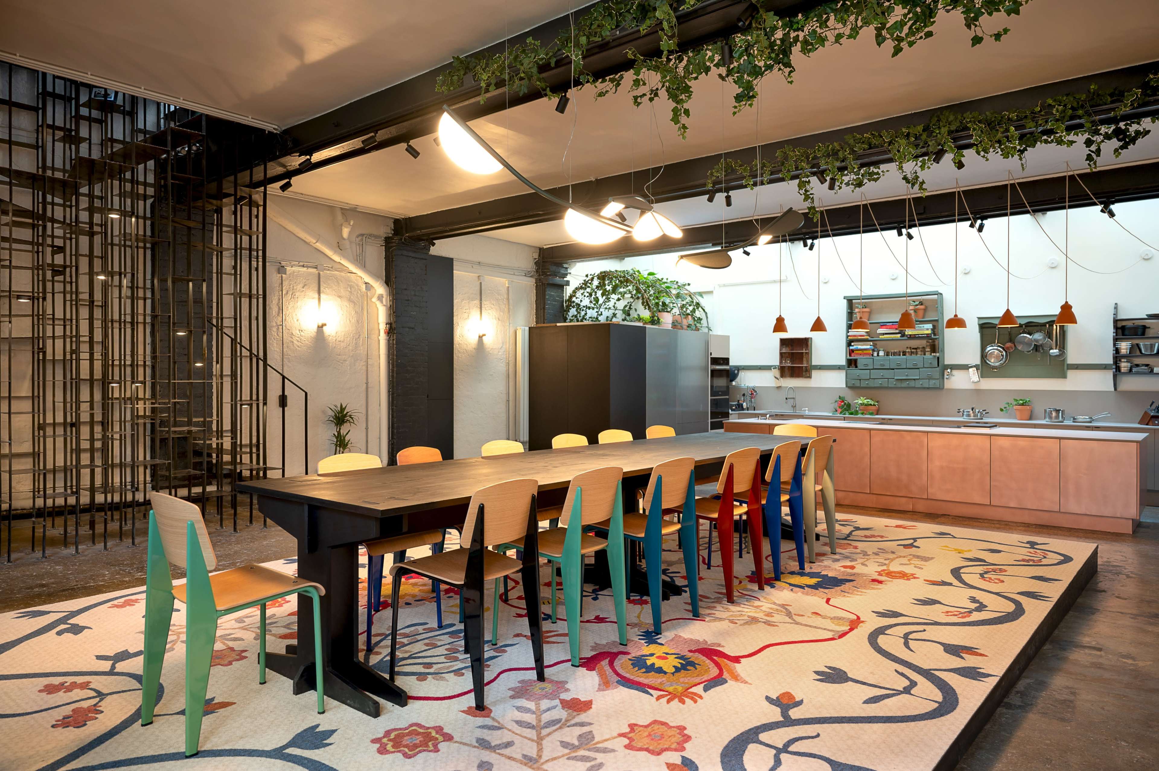 A modern, open dining area features a long table surrounded by colorful chairs, with a kitchen in the background and decorative plants hanging from the ceiling.