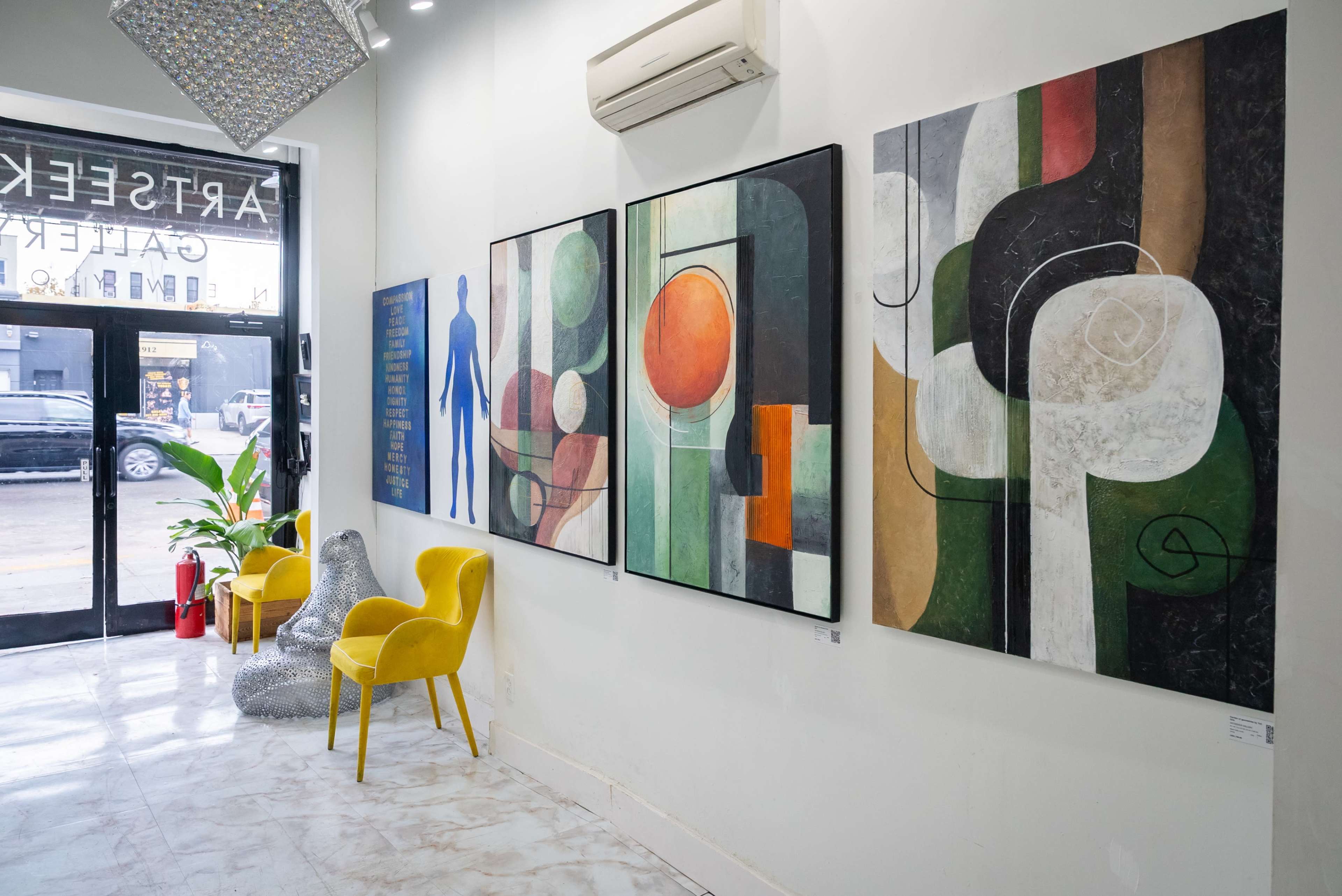 The image shows the interior of an art gallery featuring abstract paintings on the wall, a yellow chair, and a potted plant near a glass door.