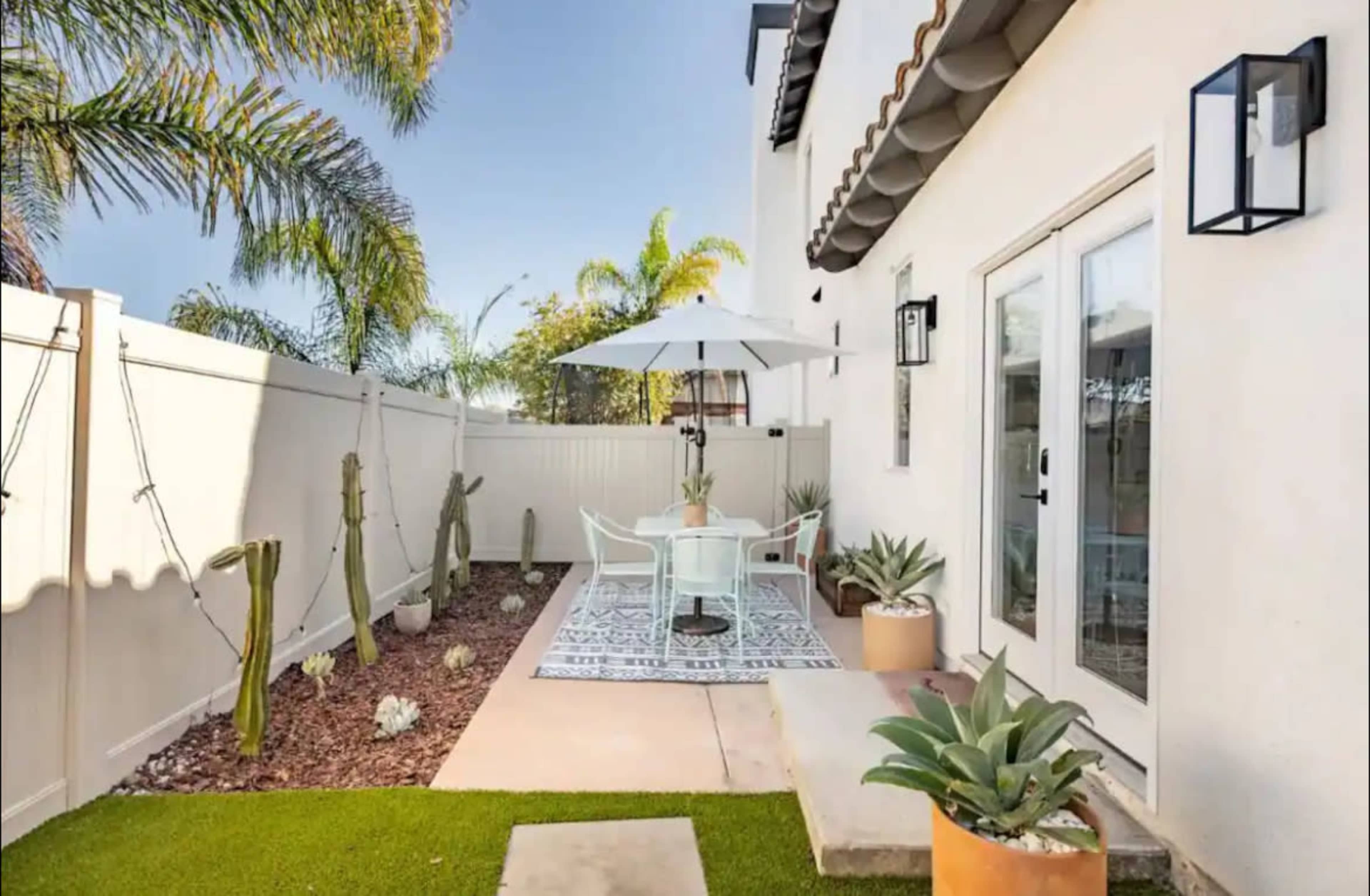 A small outdoor patio features a table with chairs, an umbrella, and potted plants alongside a decorative rug and a garden area with cacti.