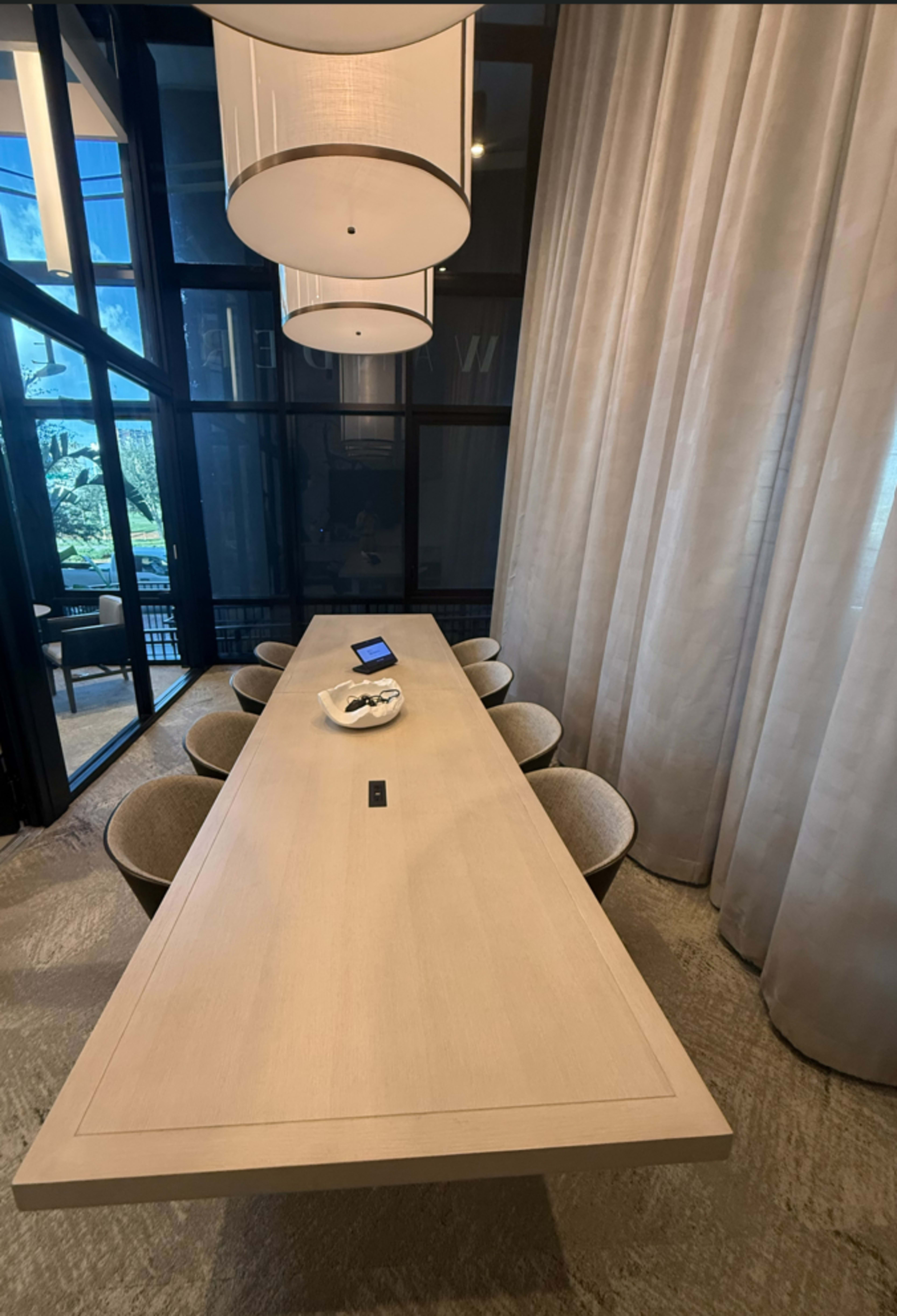 A large, modern conference table surrounded by upholstered chairs is illuminated by pendant lighting in a well-lit meeting room.