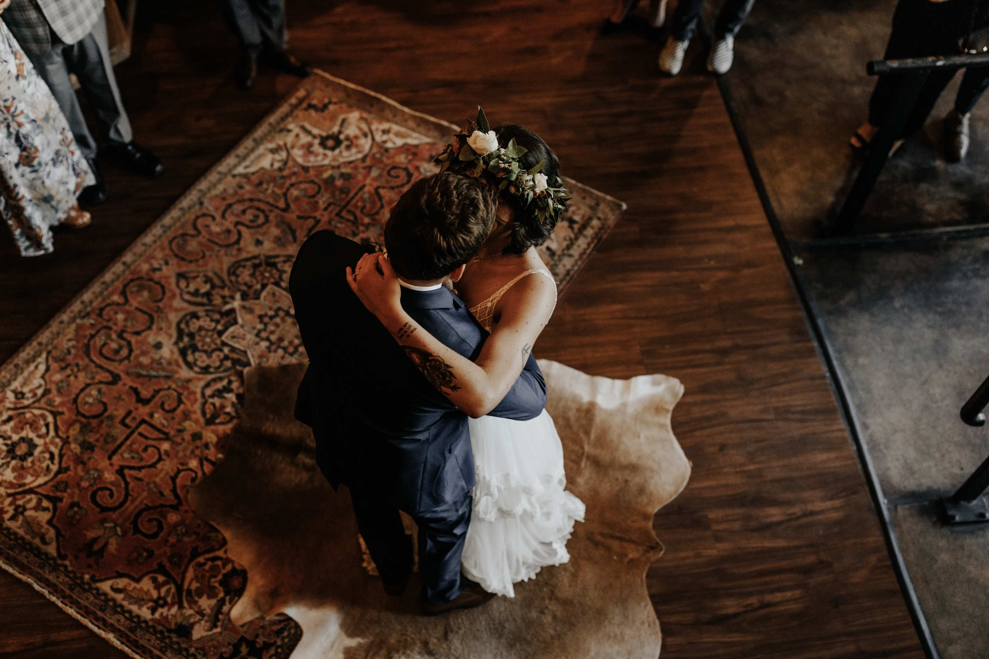 A couple dances closely on a patterned area rug in a festive indoor setting, surrounded by guests.