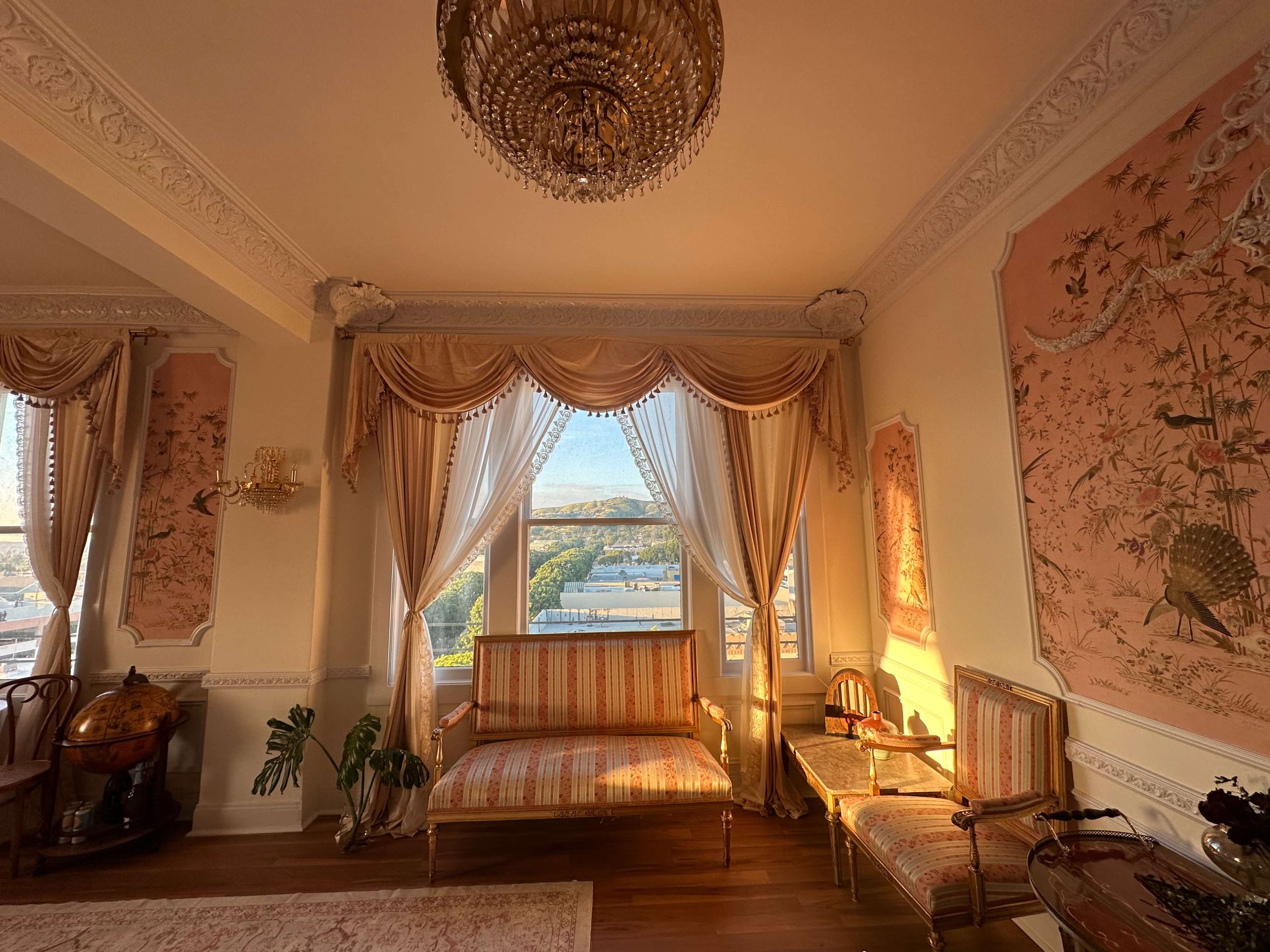 The image shows a stylishly decorated room with ornate furnishings, a large window allowing sunlight in, and pink floral wall murals.
