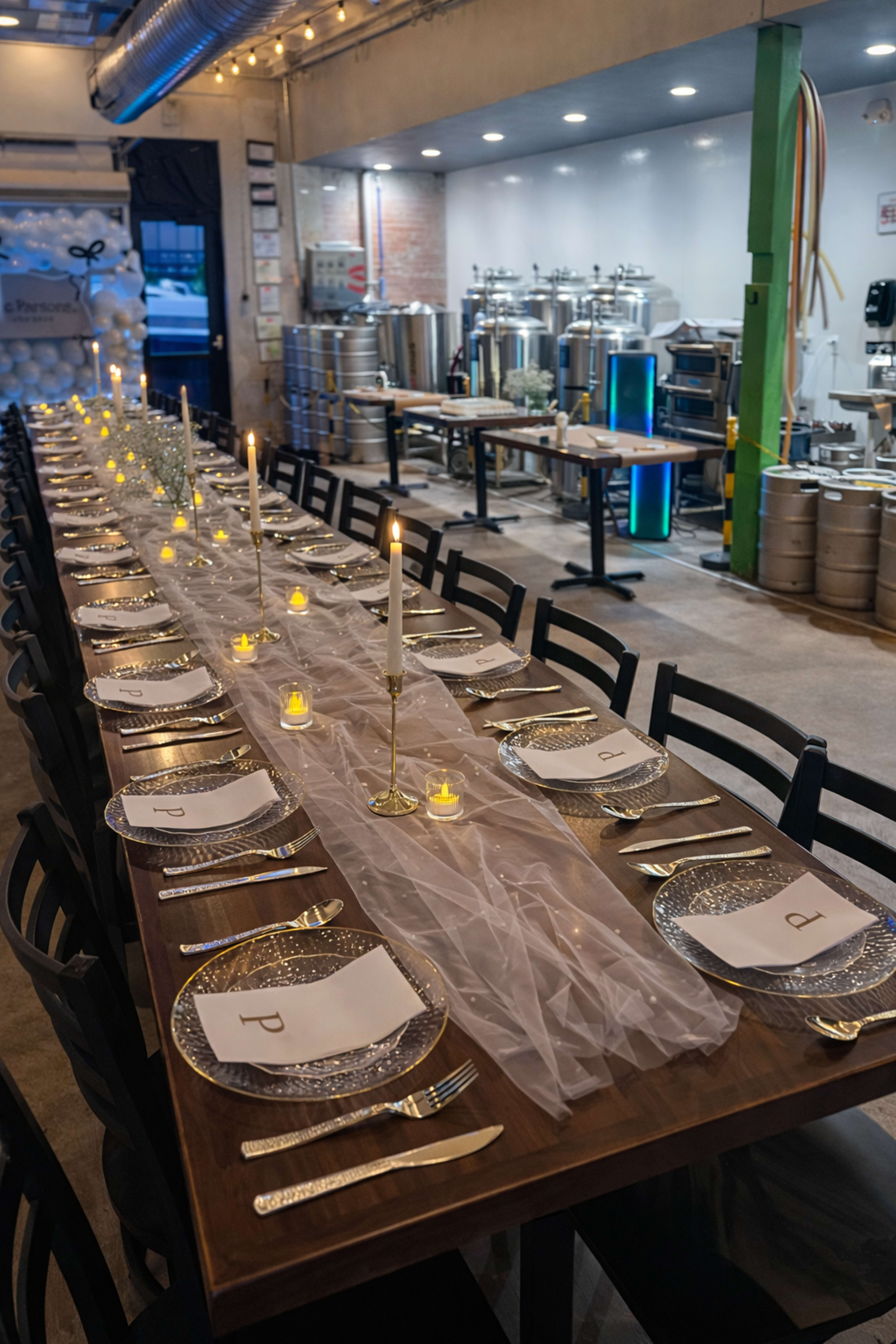 A long dining table is set with candles, elegant place settings, and a backdrop of brewing equipment in a well-lit venue.