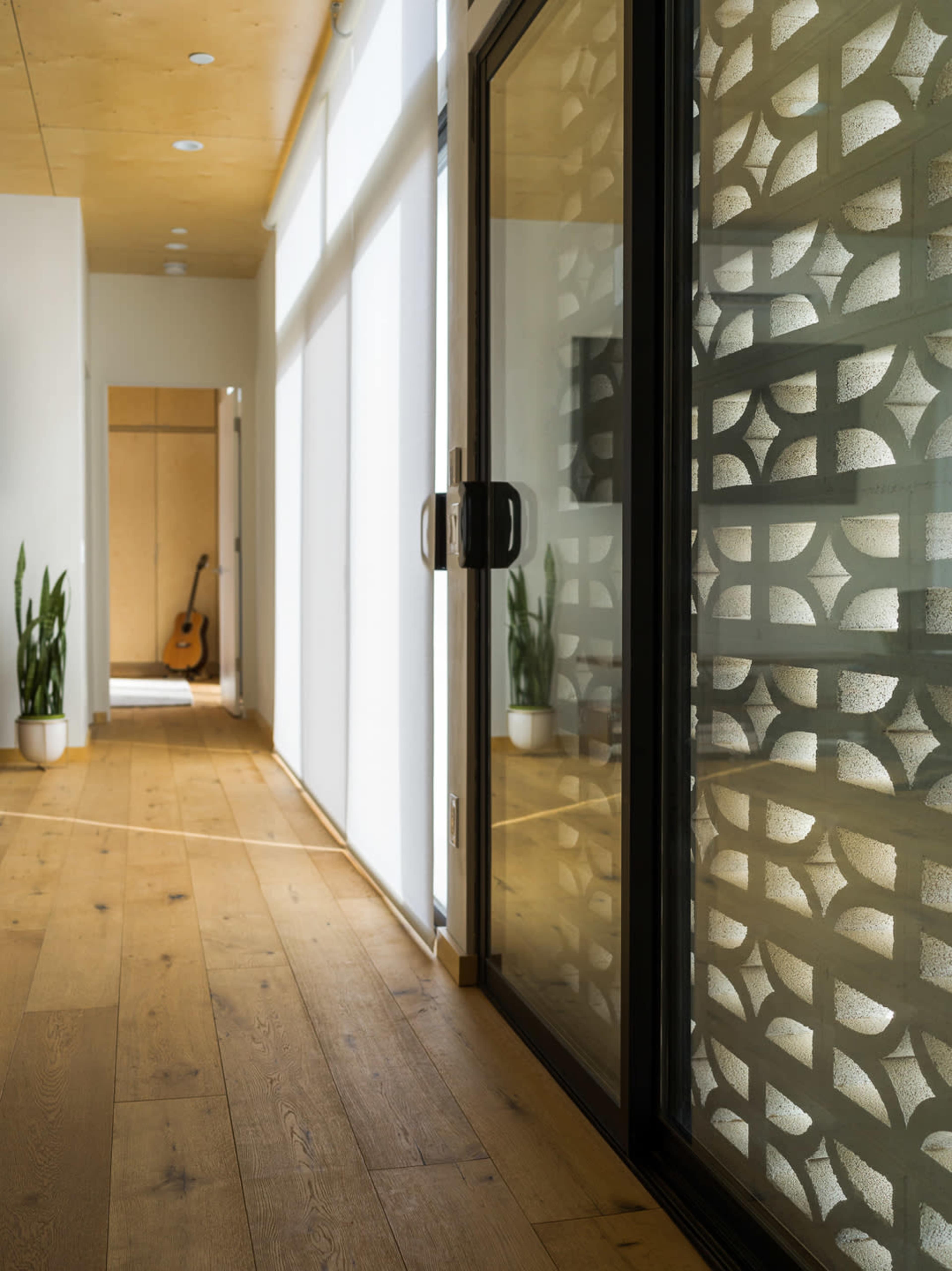 A hallway features large glass doors with a patterned design, wooden flooring, and a glimpse of a room with a guitar in the background.