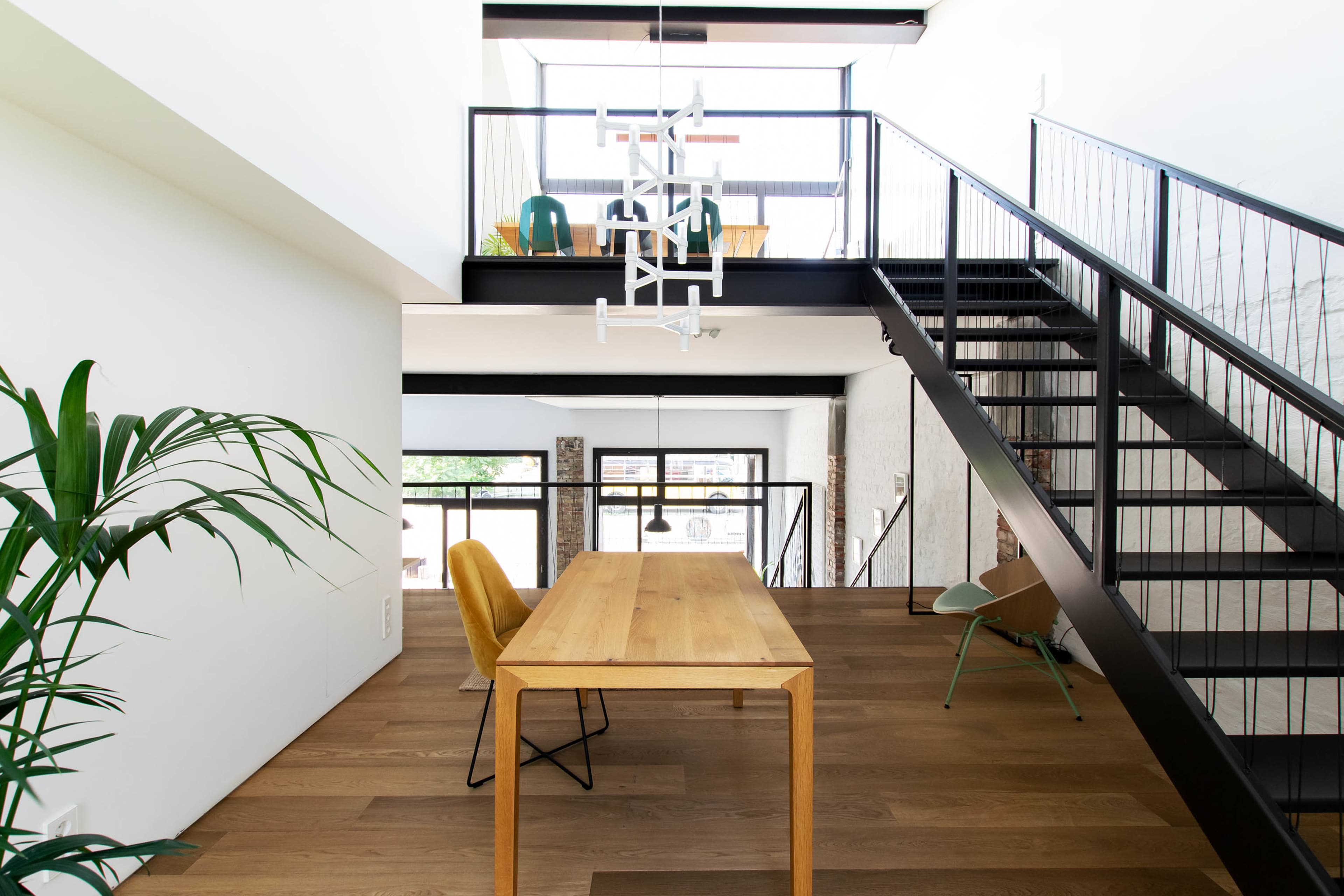 A modern interior space featuring a wooden dining table, black metal staircase, and large windows allowing natural light to enter.