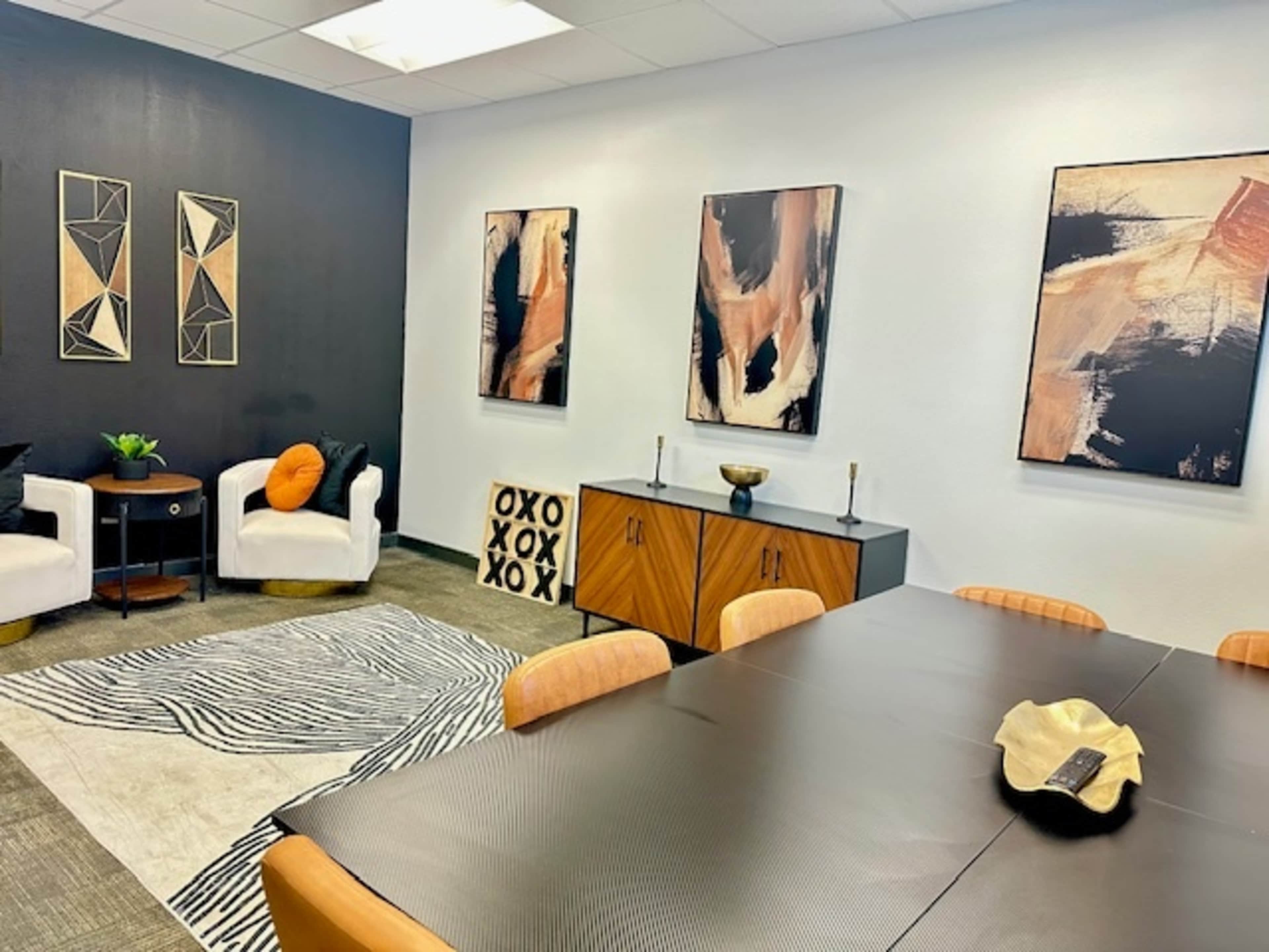 The image shows a modern meeting room adorned with abstract art, a dark accent wall, and a mixed-use seating area featuring two white chairs and a small table.