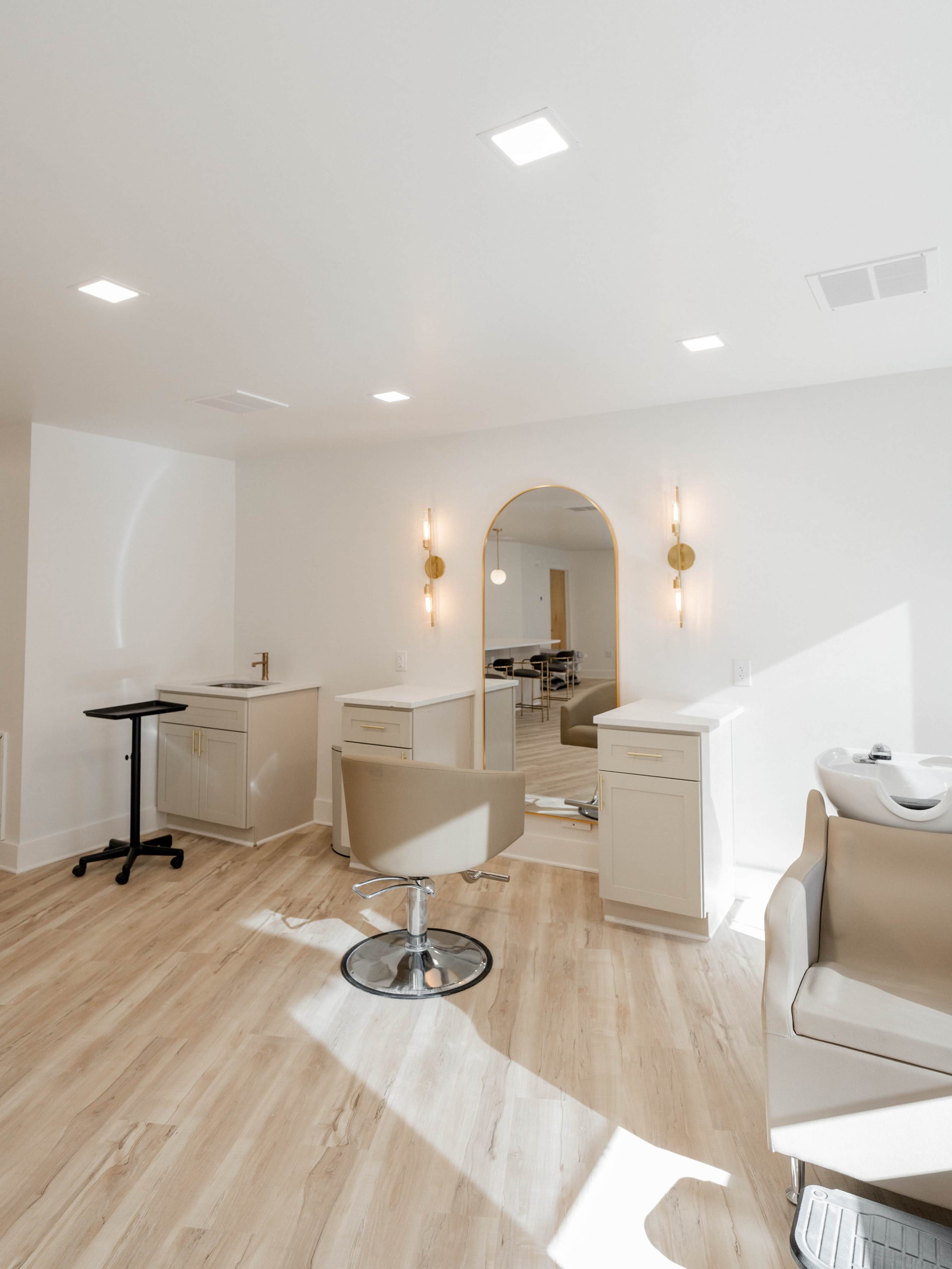 A modern hair salon features two styling stations with mirrors, a shampoo station, and light-colored wooden flooring.