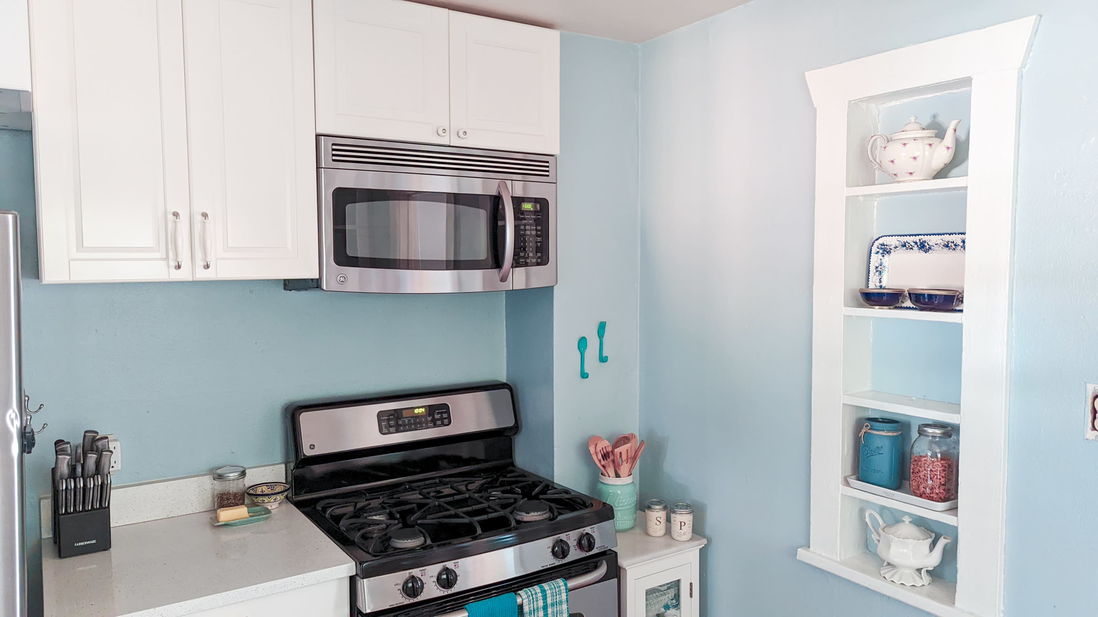 A modern kitchen features a stainless steel stove and microwave against light blue walls, with white cabinetry and shelves displaying various kitchen items.