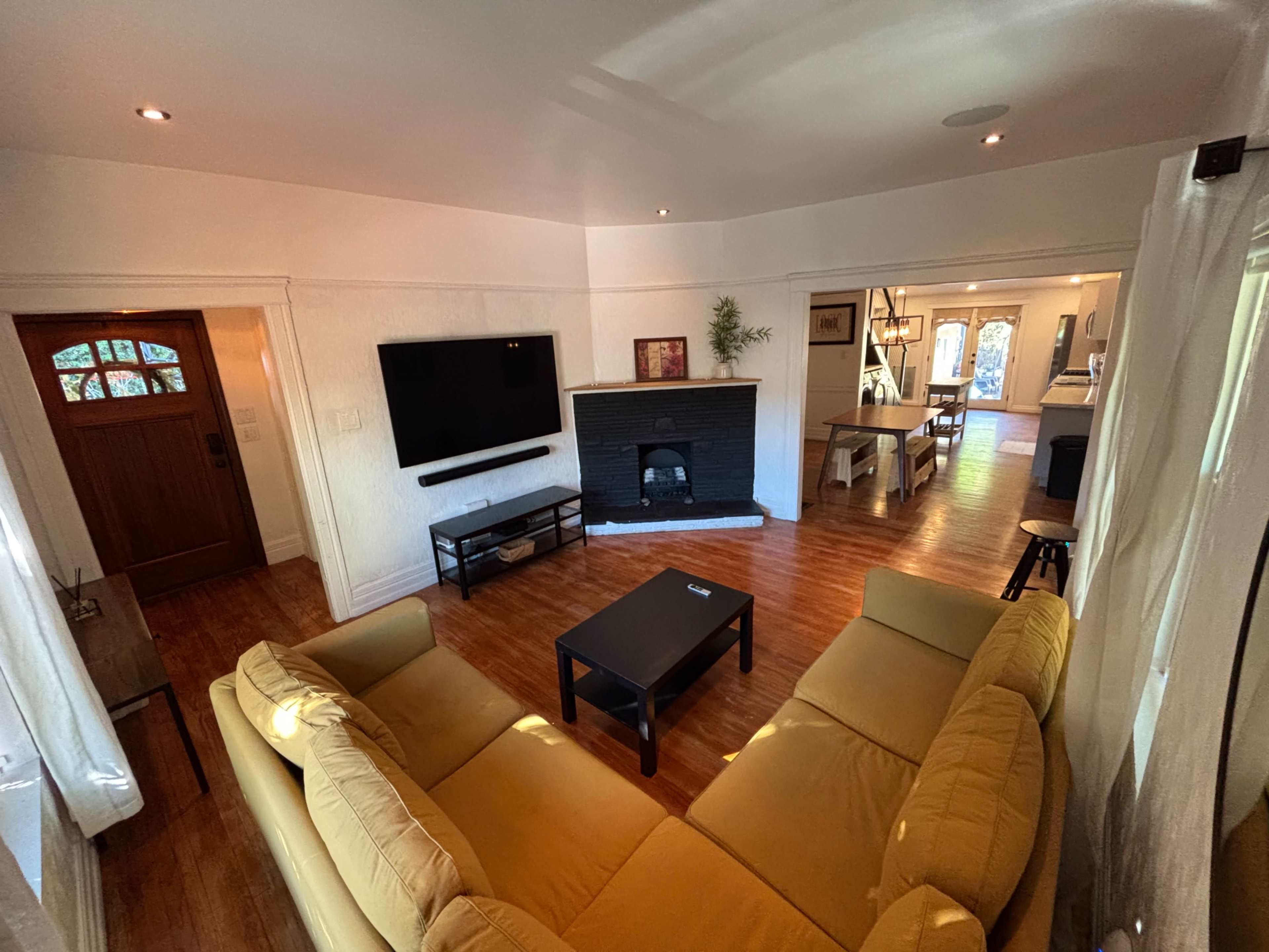 The image shows a living room with a yellow sectional sofa, a black coffee table, a wall-mounted TV, and a fireplace, adjacent to an entryway and a dining area.