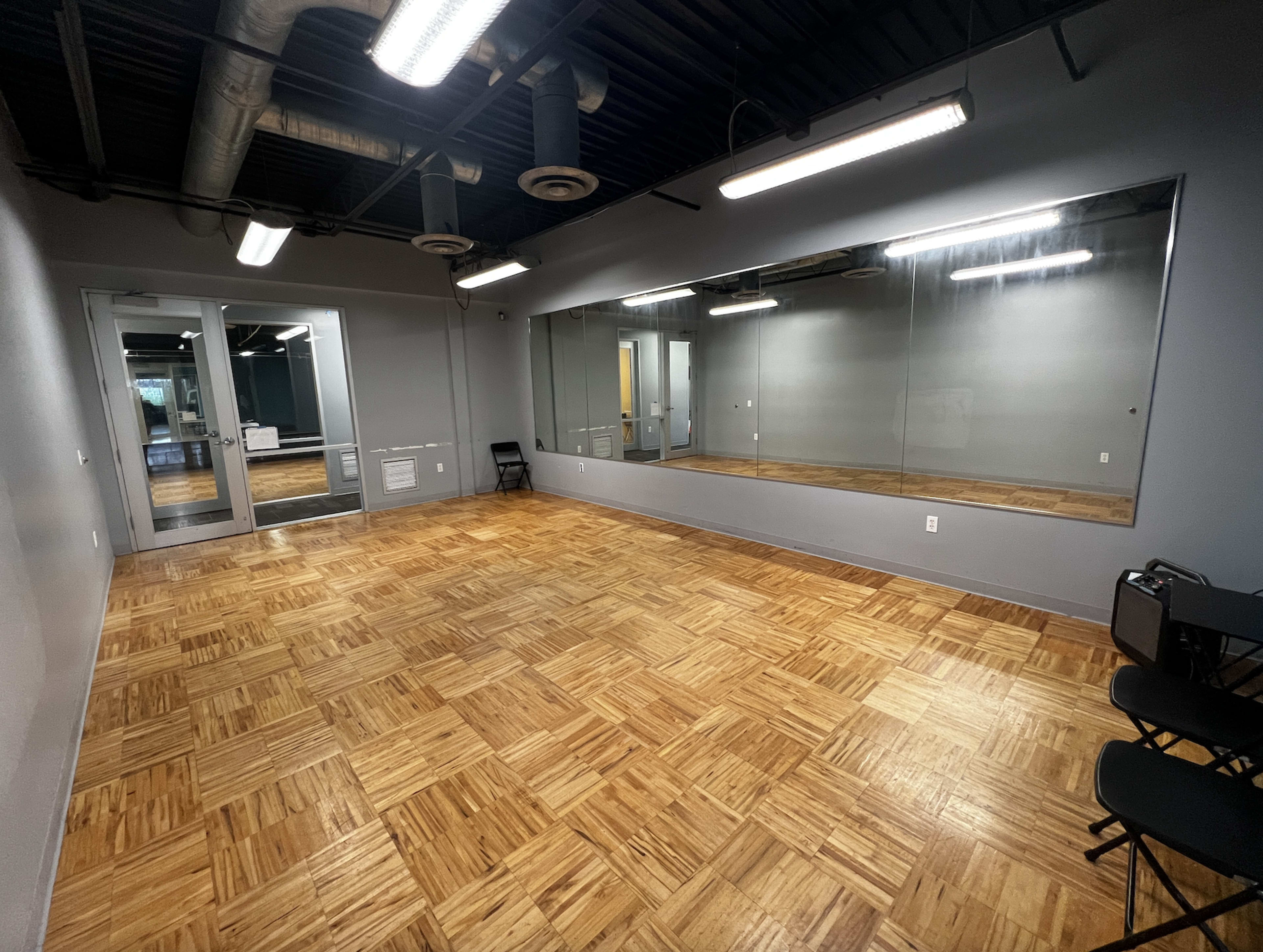 An empty room with wooden flooring, gray walls, a large mirror on one side, and chairs positioned along the opposite wall.