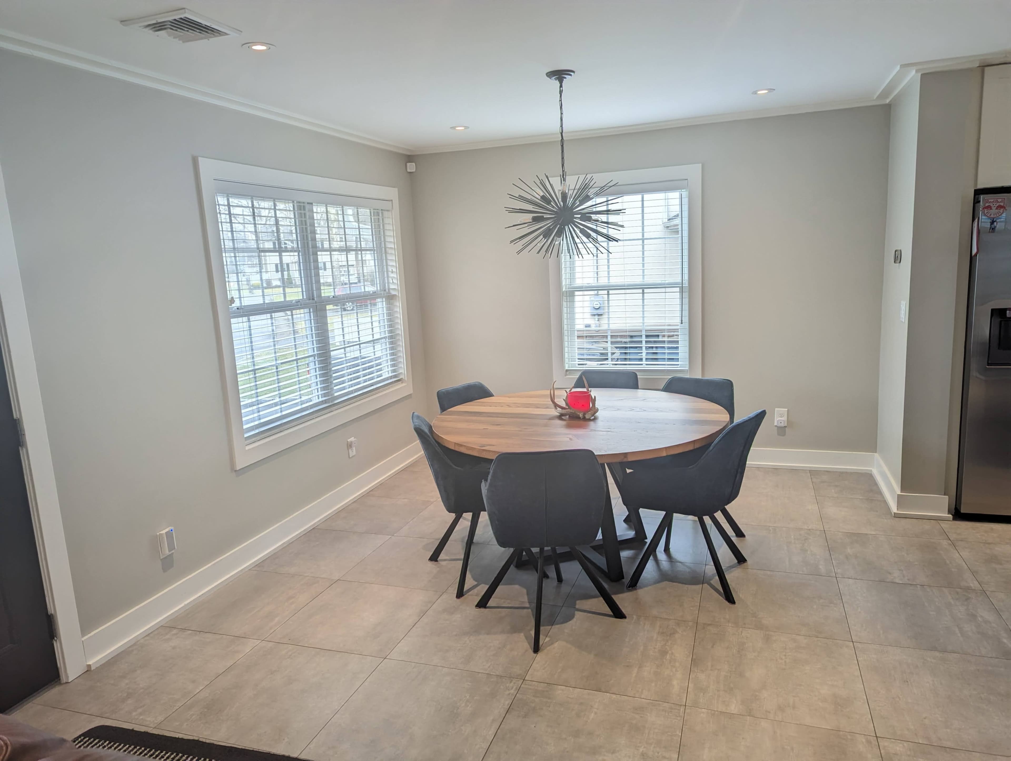 A round wooden dining table surrounded by six gray chairs is positioned in a well-lit room with white walls and large windows.