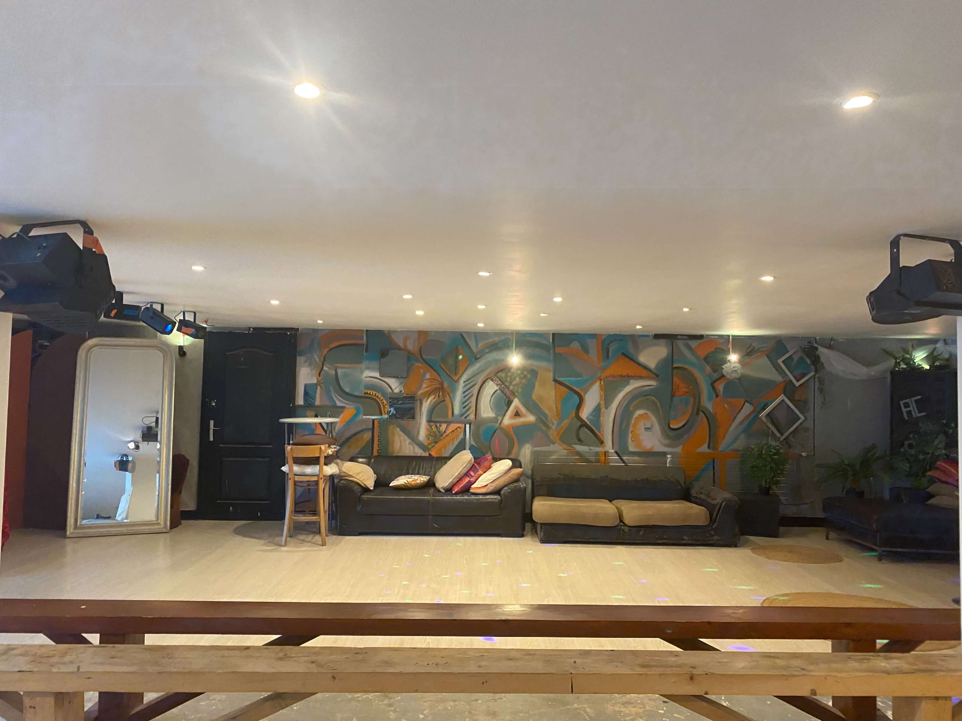 A spacious indoor area features a mural on the wall, two black sofas, a wooden bar table, and various decorative plants.