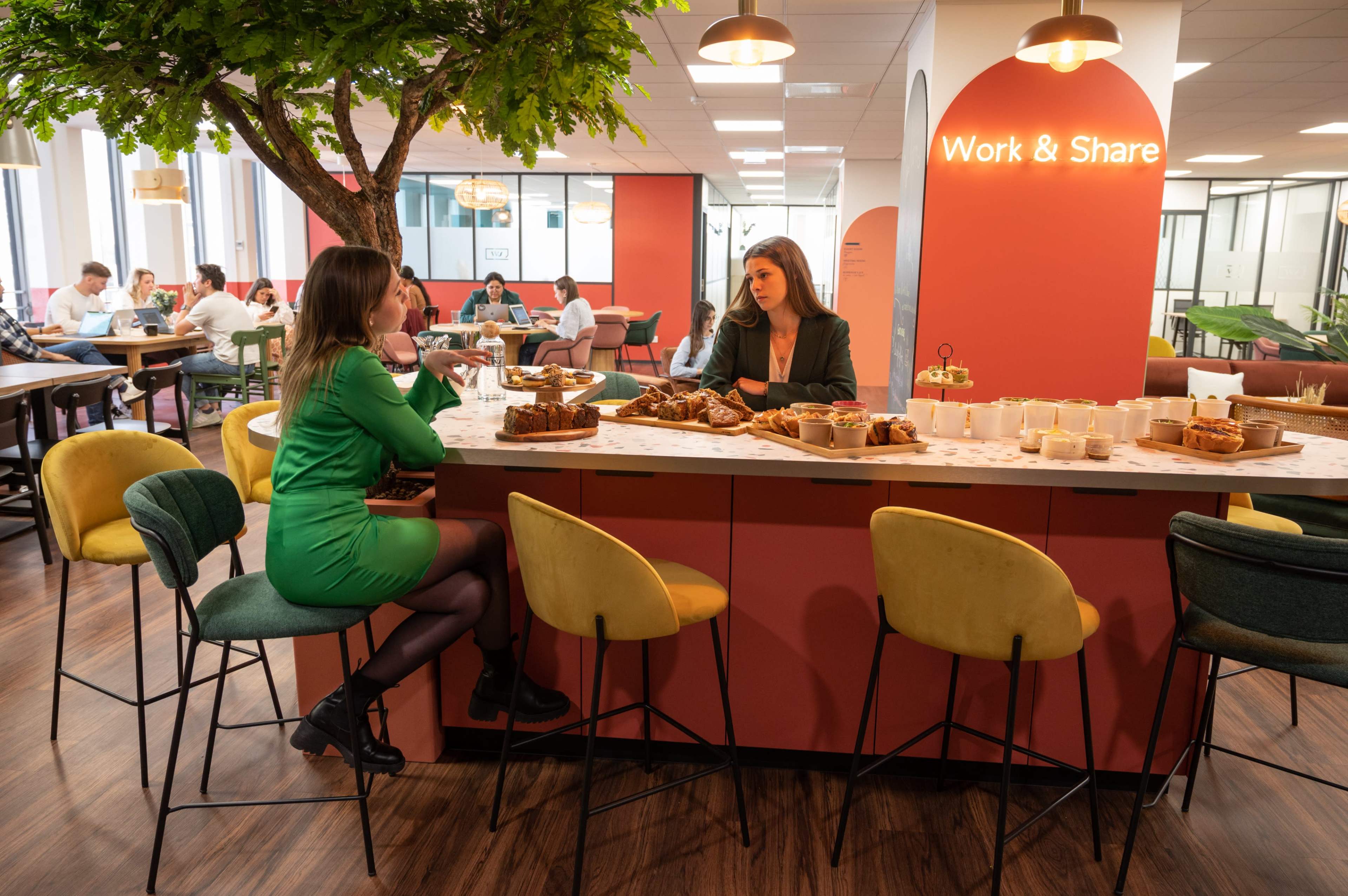 Two women sit at a circular bar in a modern office space, surrounded by greenery and pastries, while others work at tables in the background.