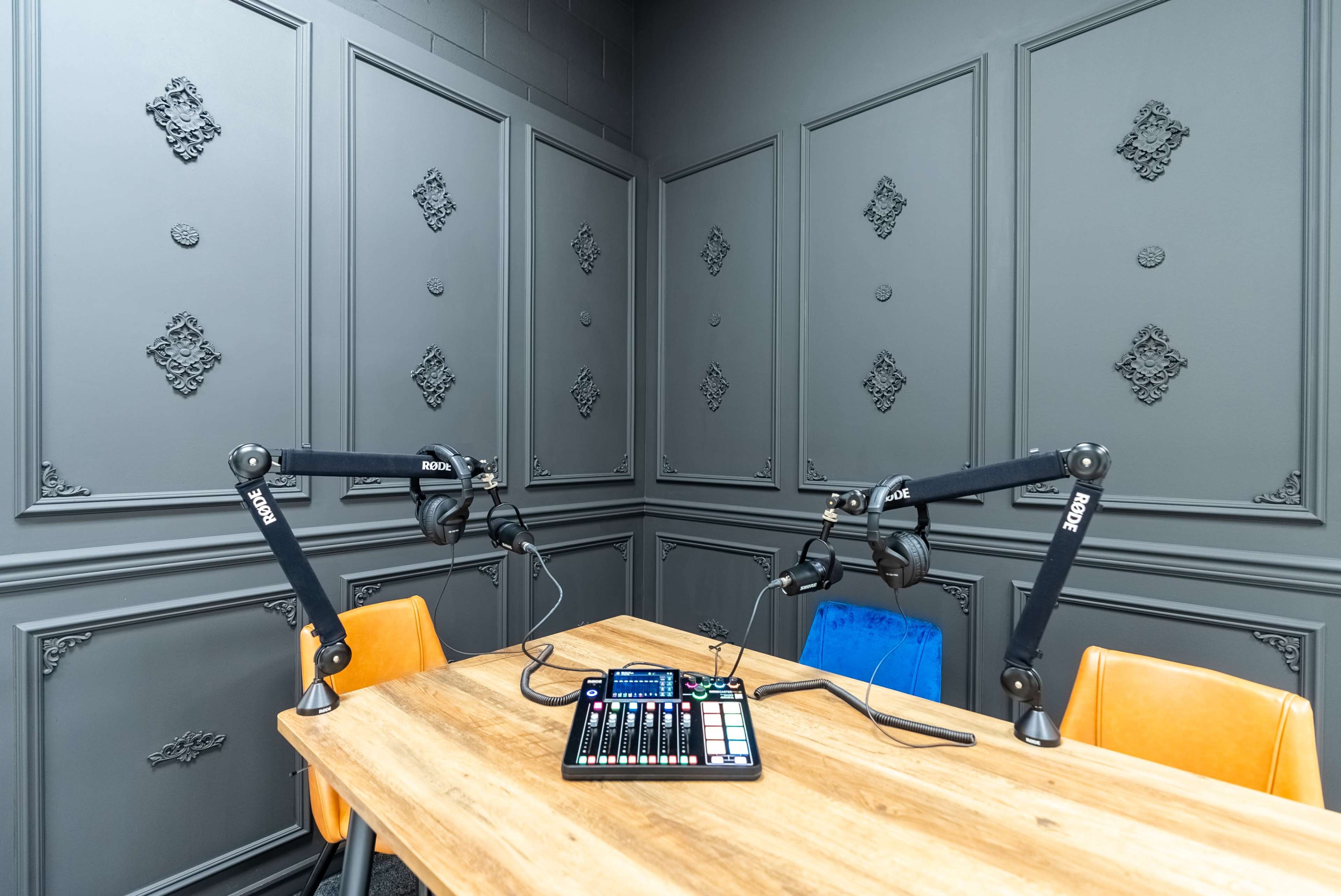 A recording studio features a wooden table with audio equipment and two microphone stands against textured gray walls.