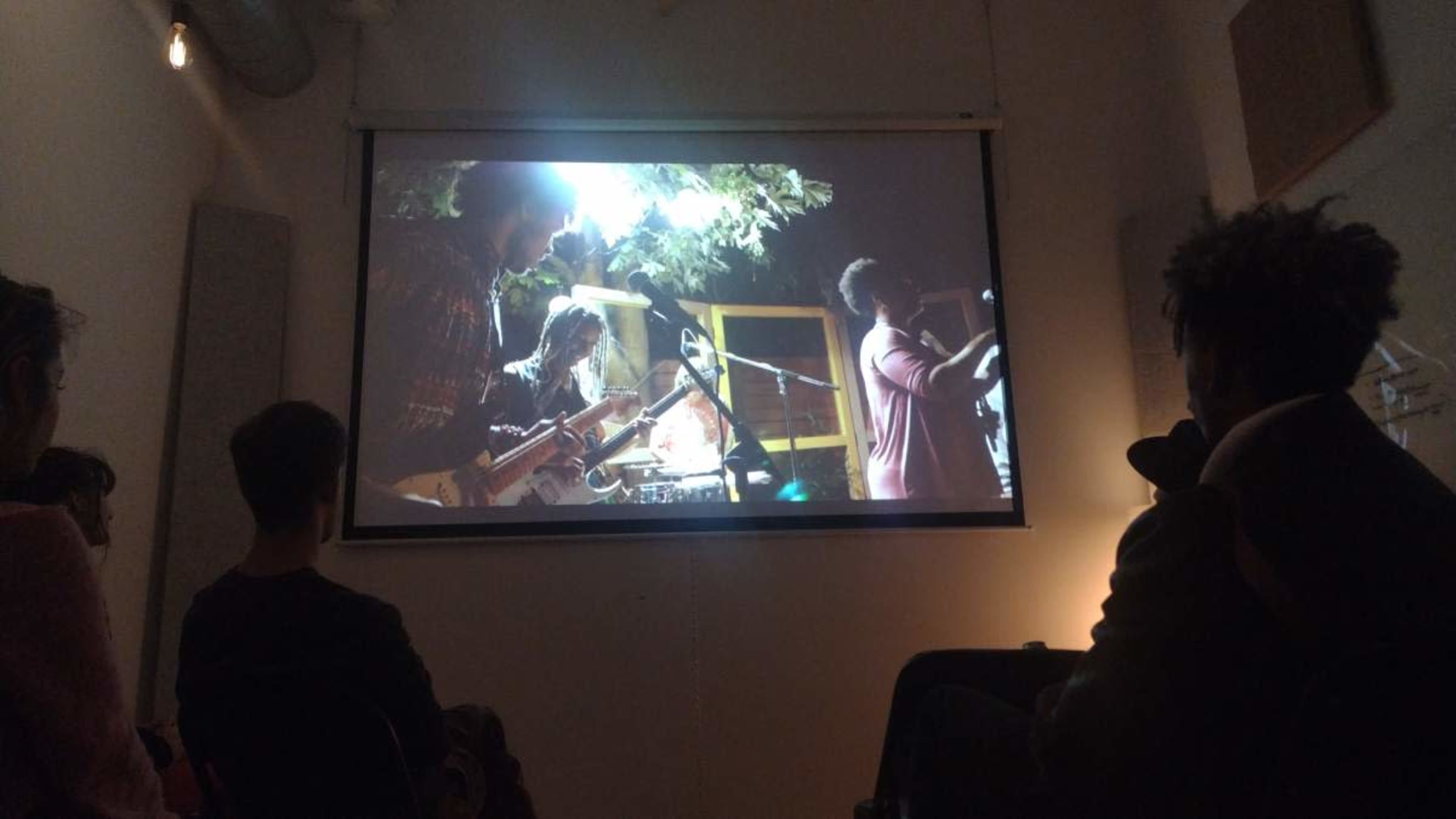 A group of people is watching a band perform on a large screen in a dimly lit room.