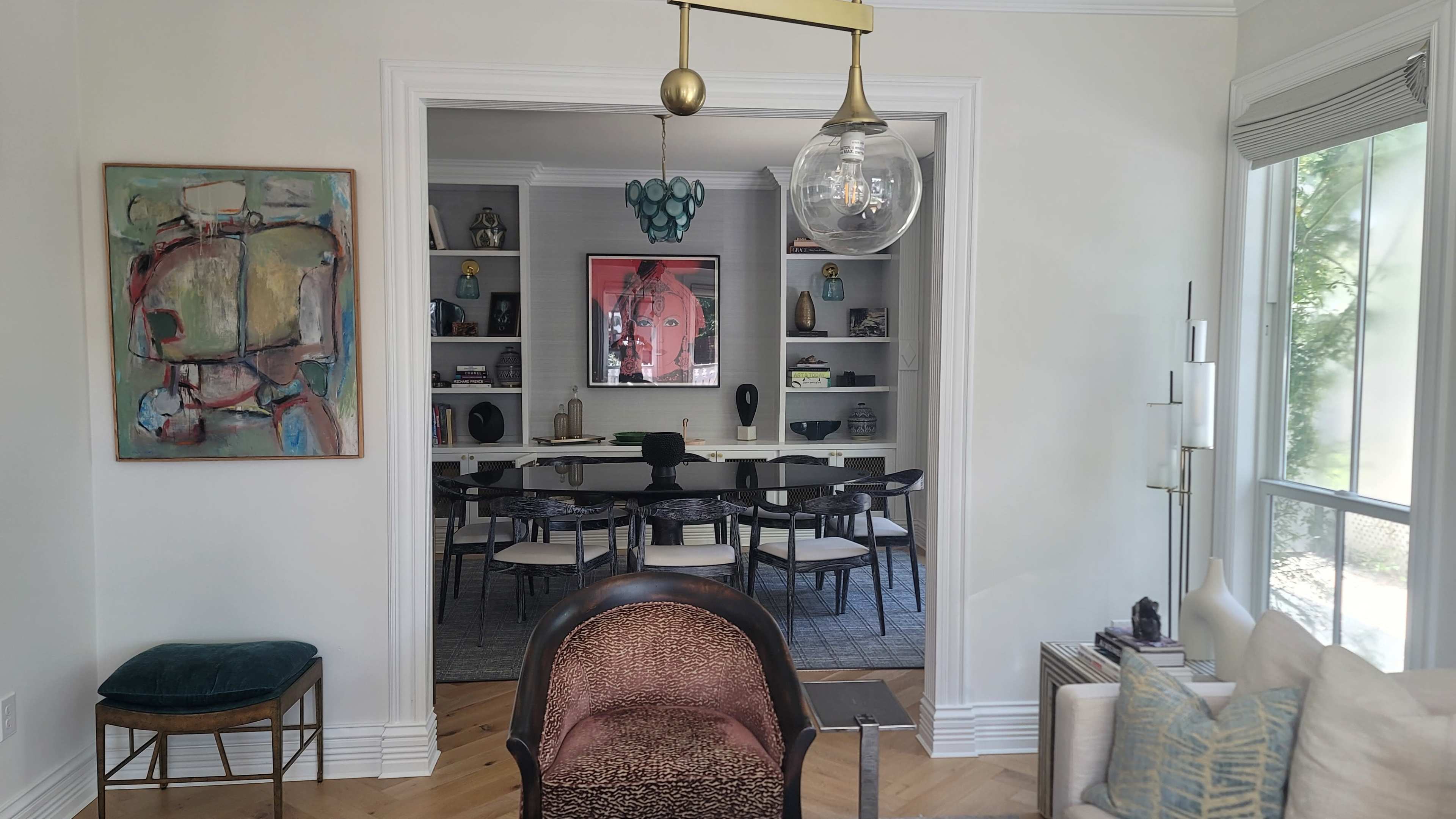 The image shows a modern living space with a view through an archway into a dining area featuring a long table surrounded by chairs and shelves displaying various decorative objects.