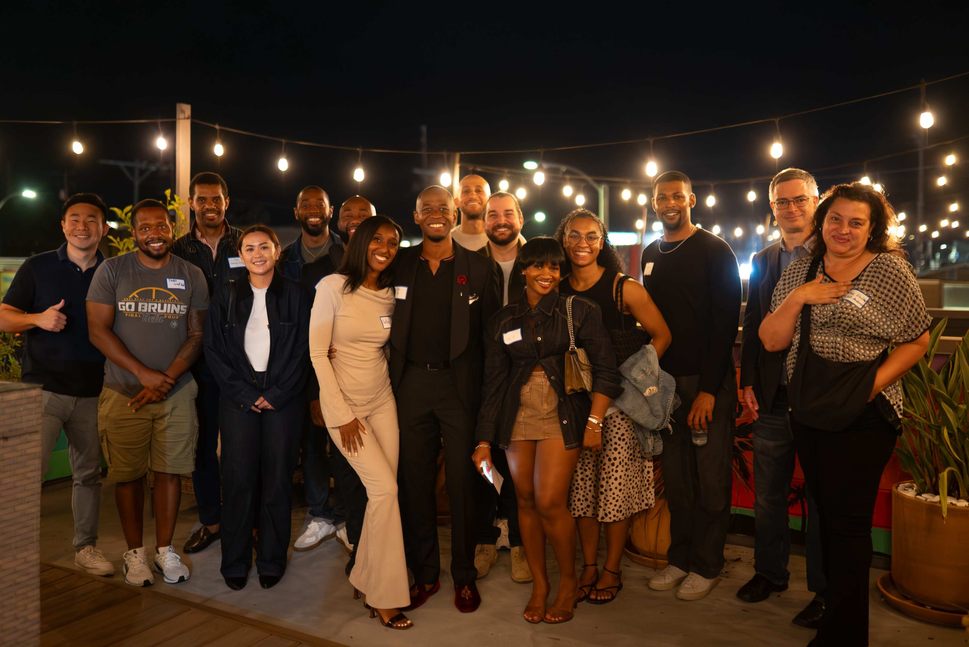 A group of diverse individuals stands together, smiling and posing for a photograph at an outdoor evening event decorated with string lights.