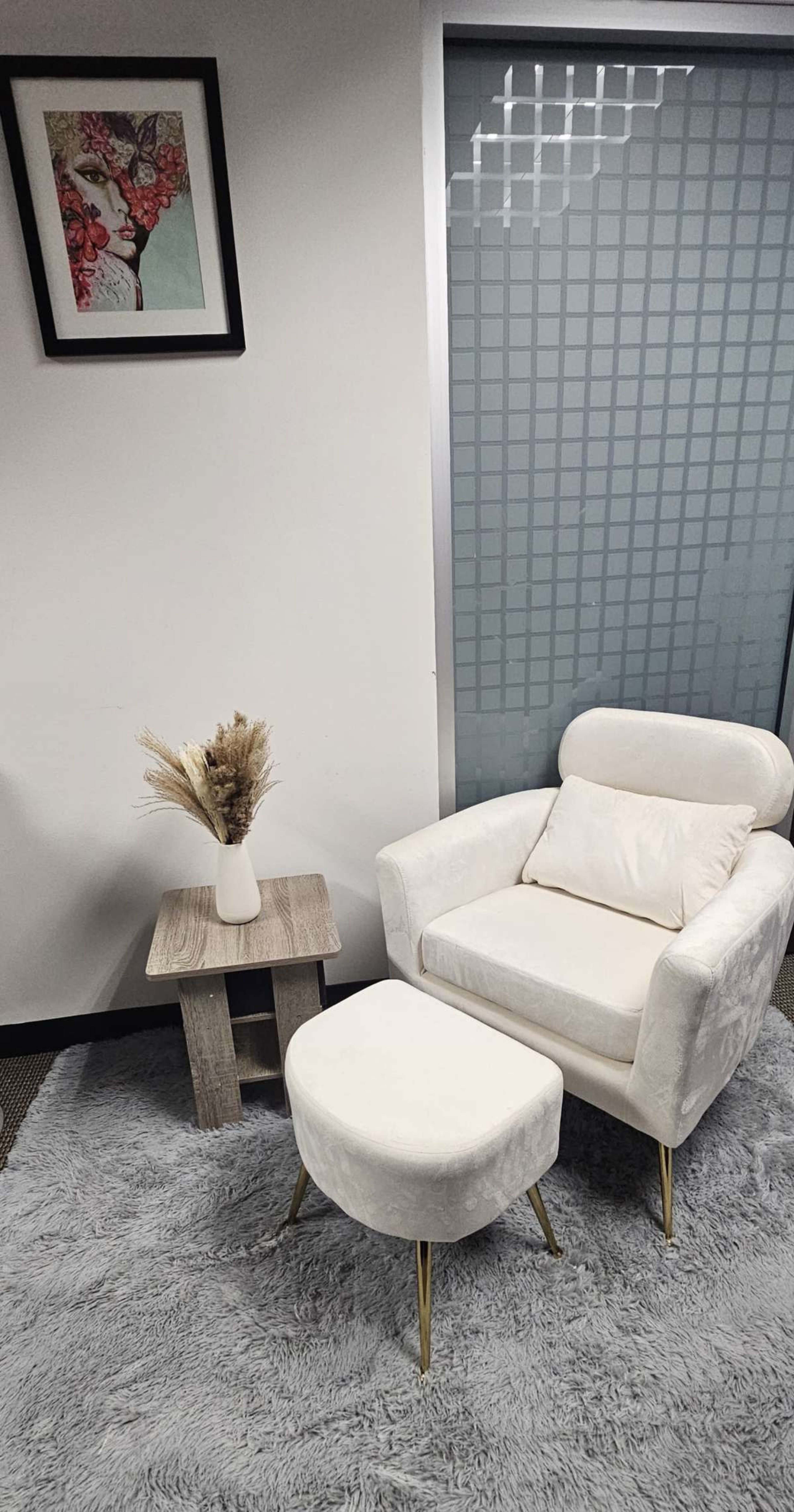 A white armchair with a matching ottoman is positioned next to a small wooden side table on a gray rug, with a framed artwork hanging on the wall above.
