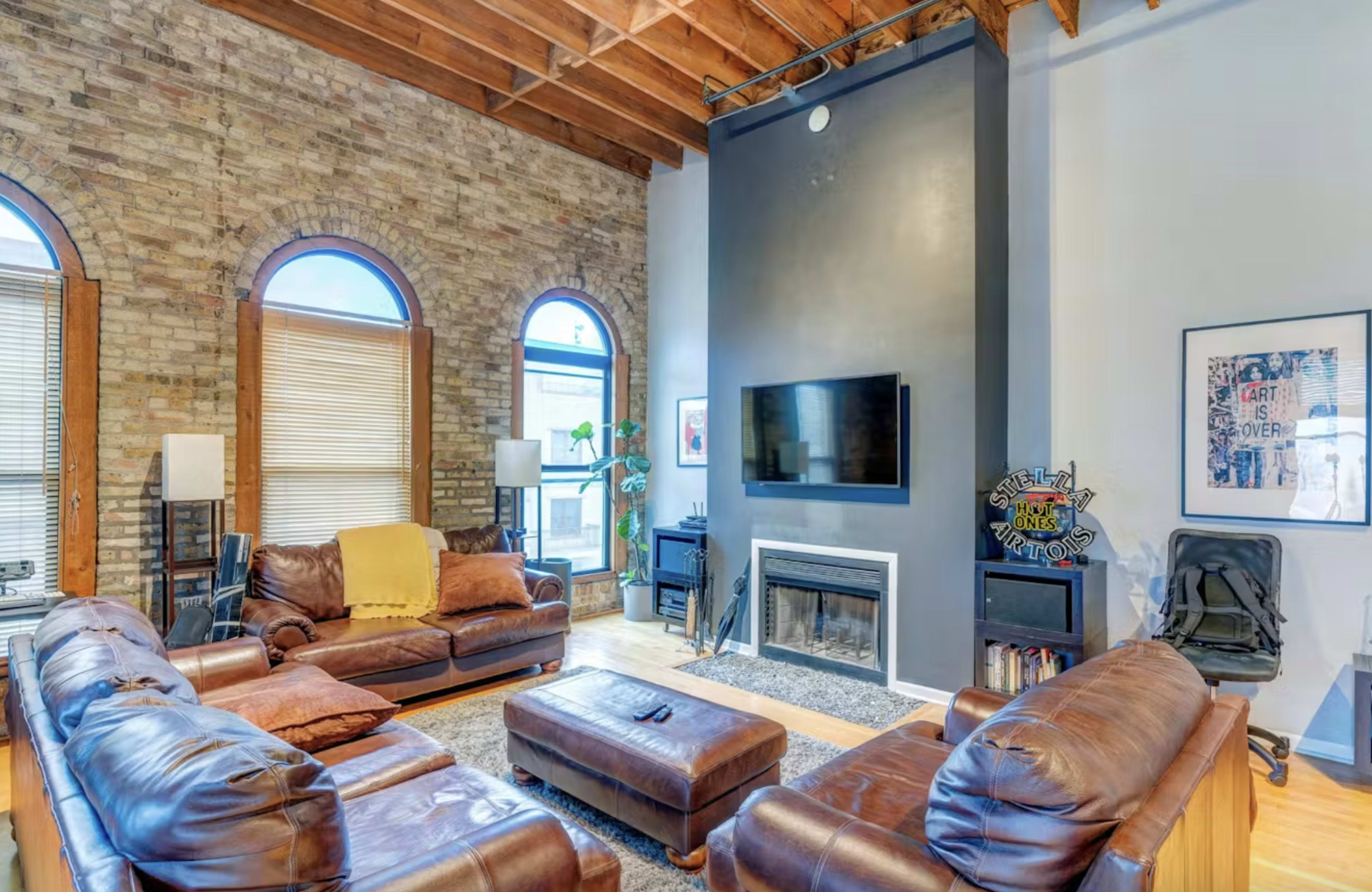 The living room features exposed brick walls, leather sofas arranged around a rug, and a wall-mounted television above a fireplace.