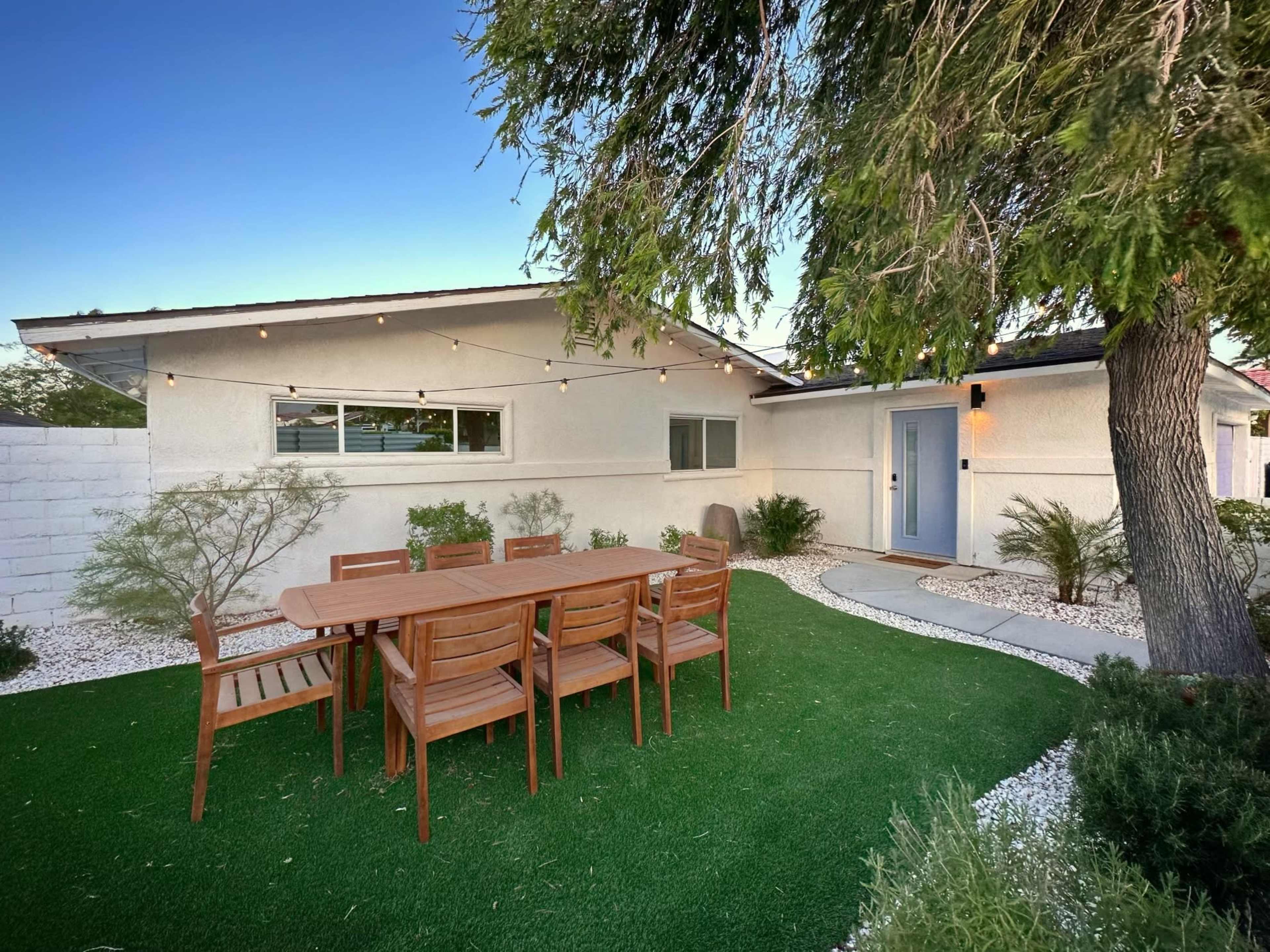A wooden dining table with eight chairs is set on artificial grass beside a modern house surrounded by white gravel and shrubs.