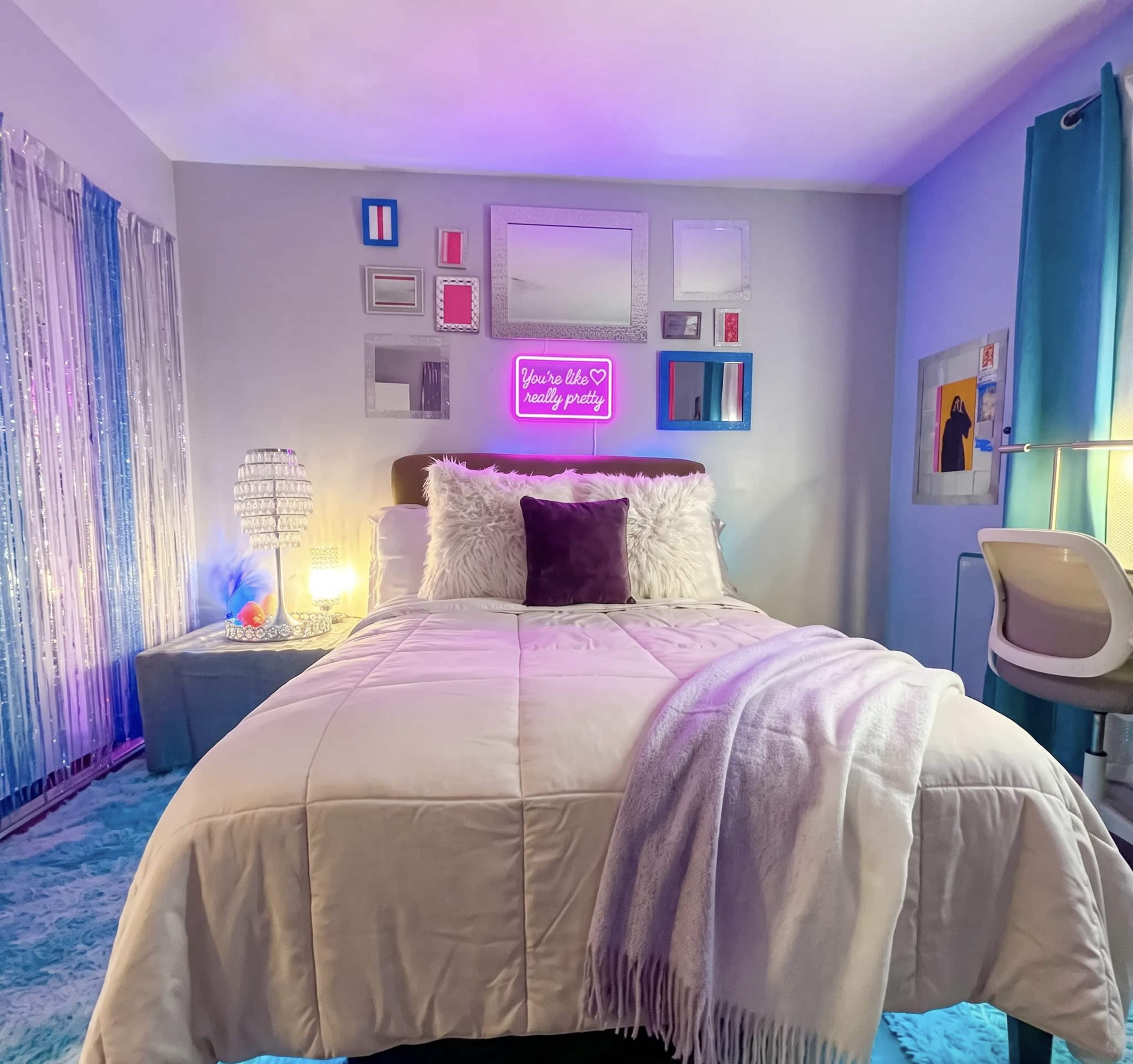 The image shows a neatly arranged bedroom with a bed covered in a white comforter, decorative pillows, and colorful wall art illuminated by a neon sign.