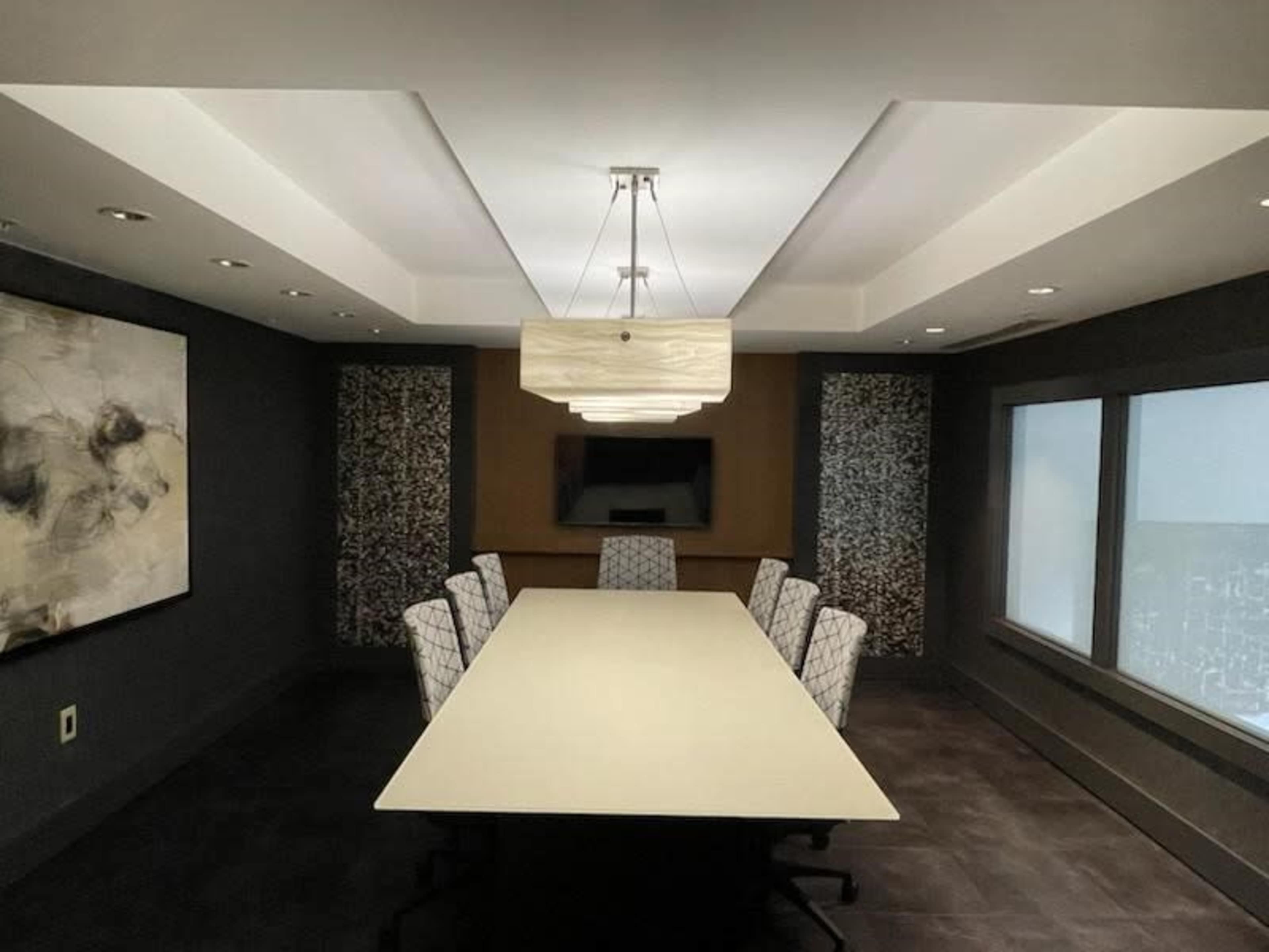 A modern conference room with a long table and six upholstered chairs, illuminated by a chandelier, and featuring large windows and artwork on the walls.