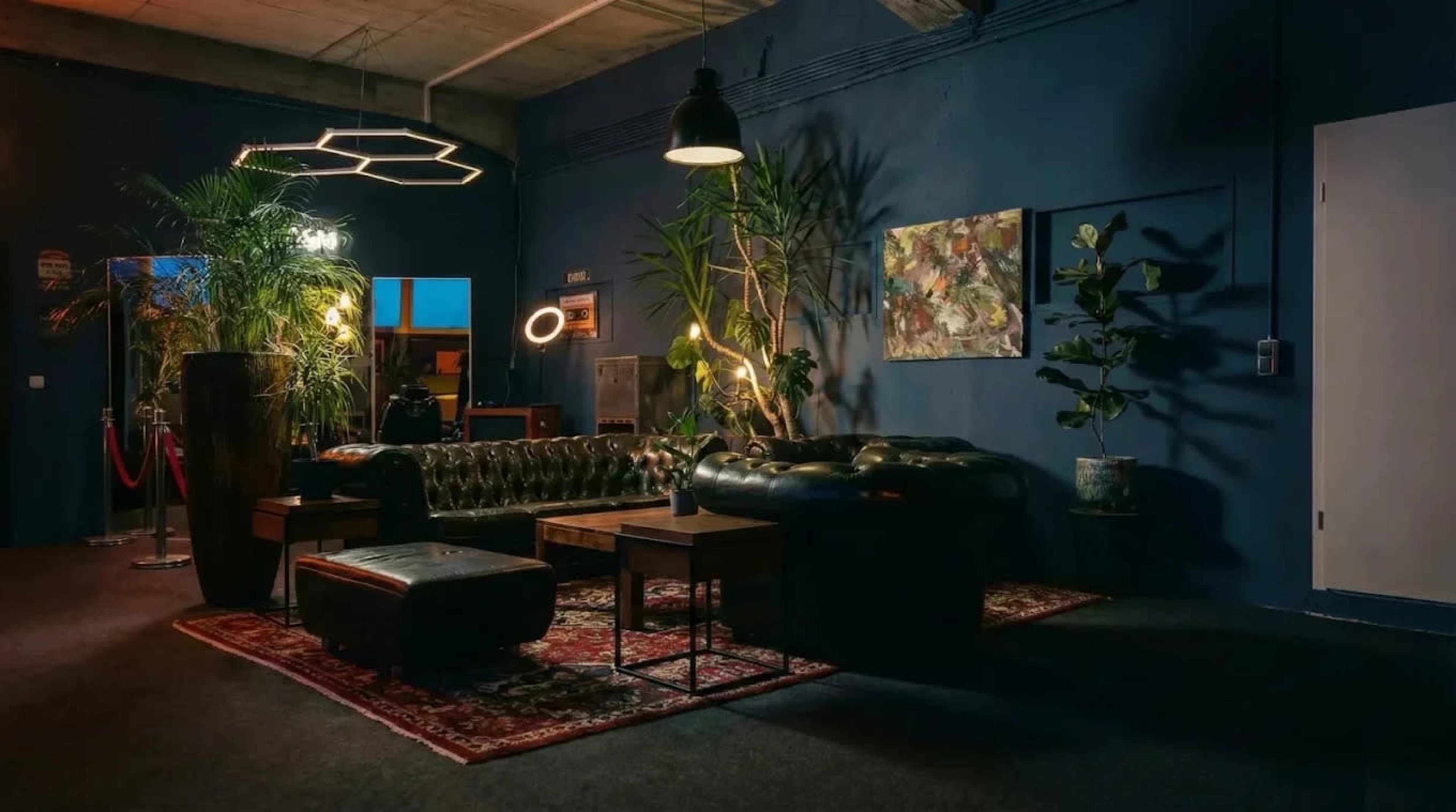 The image shows a stylish lounge area featuring a dark leather sofa, a wooden coffee table, lush plants, and warm lighting against a blue wall.