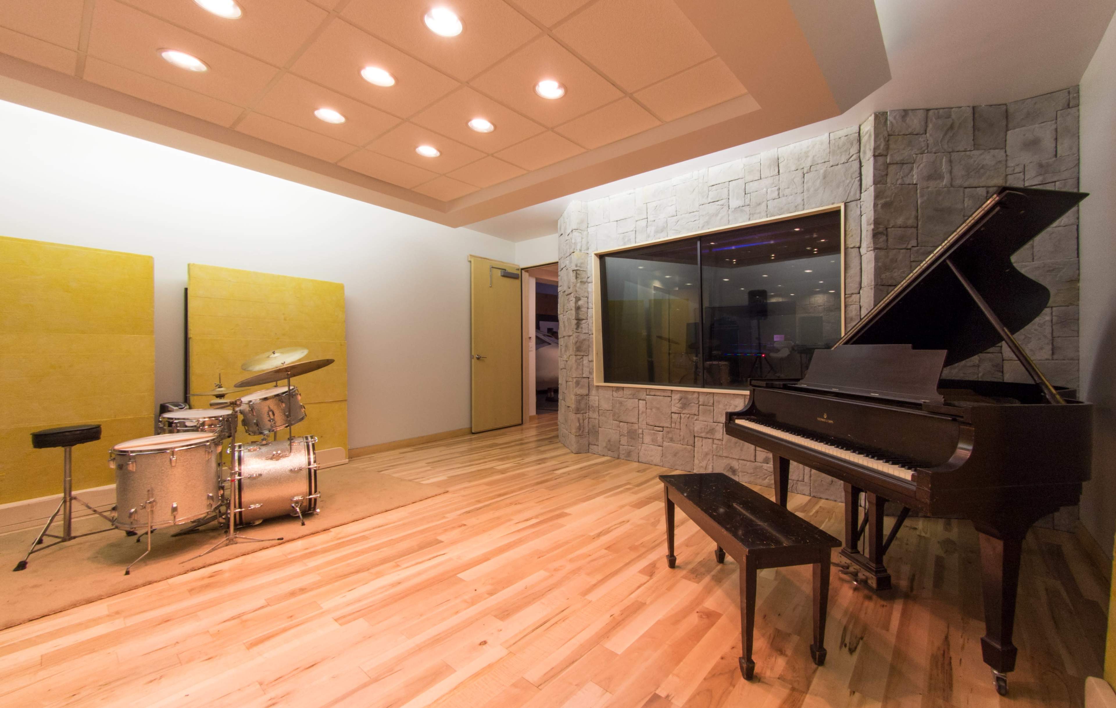 The image shows a music studio featuring a drum set, a grand piano, and a glass-enclosed recording booth.