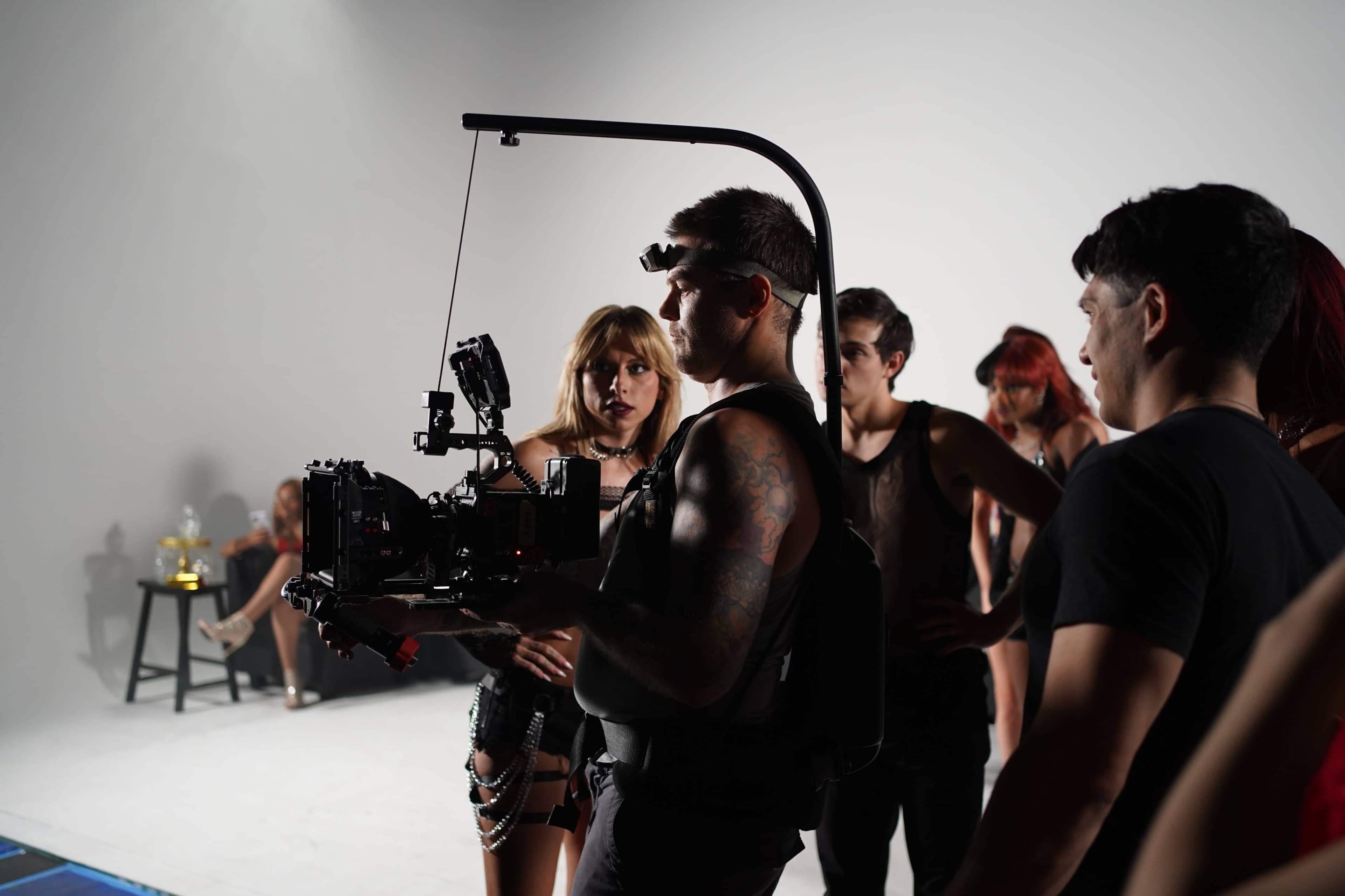A film crew is gathered on a set, with a camera operator in a harness holding a professional camera, while a female performer and several other crew members observe.