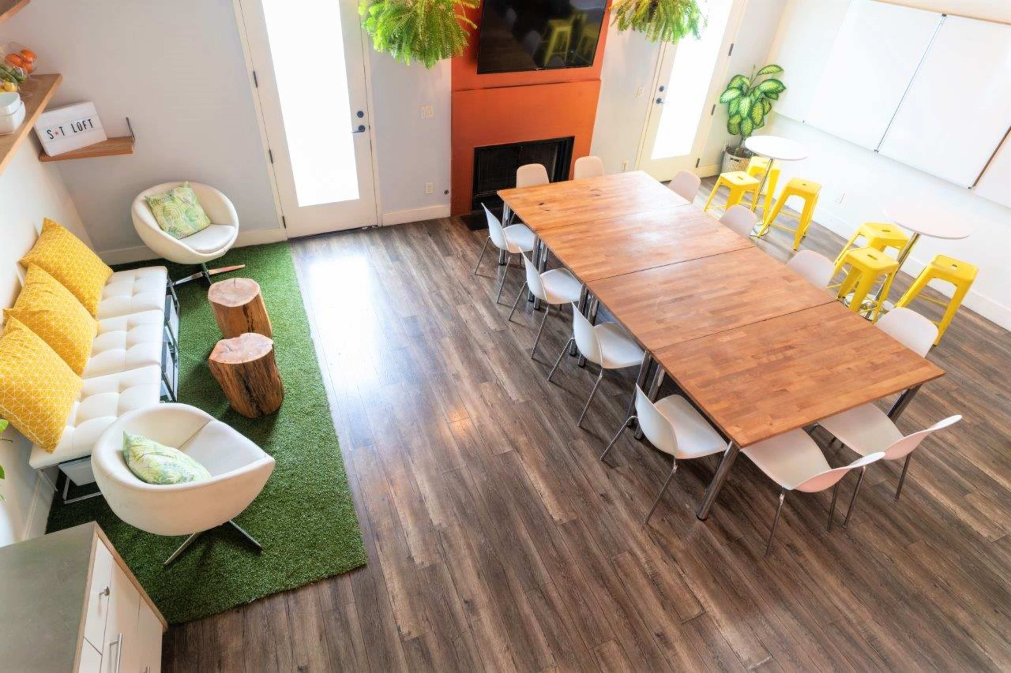 A modern meeting room with a large wooden table surrounded by white chairs, two white armchairs, and a green rug, set against a backdrop of windows and a wall-mounted television.