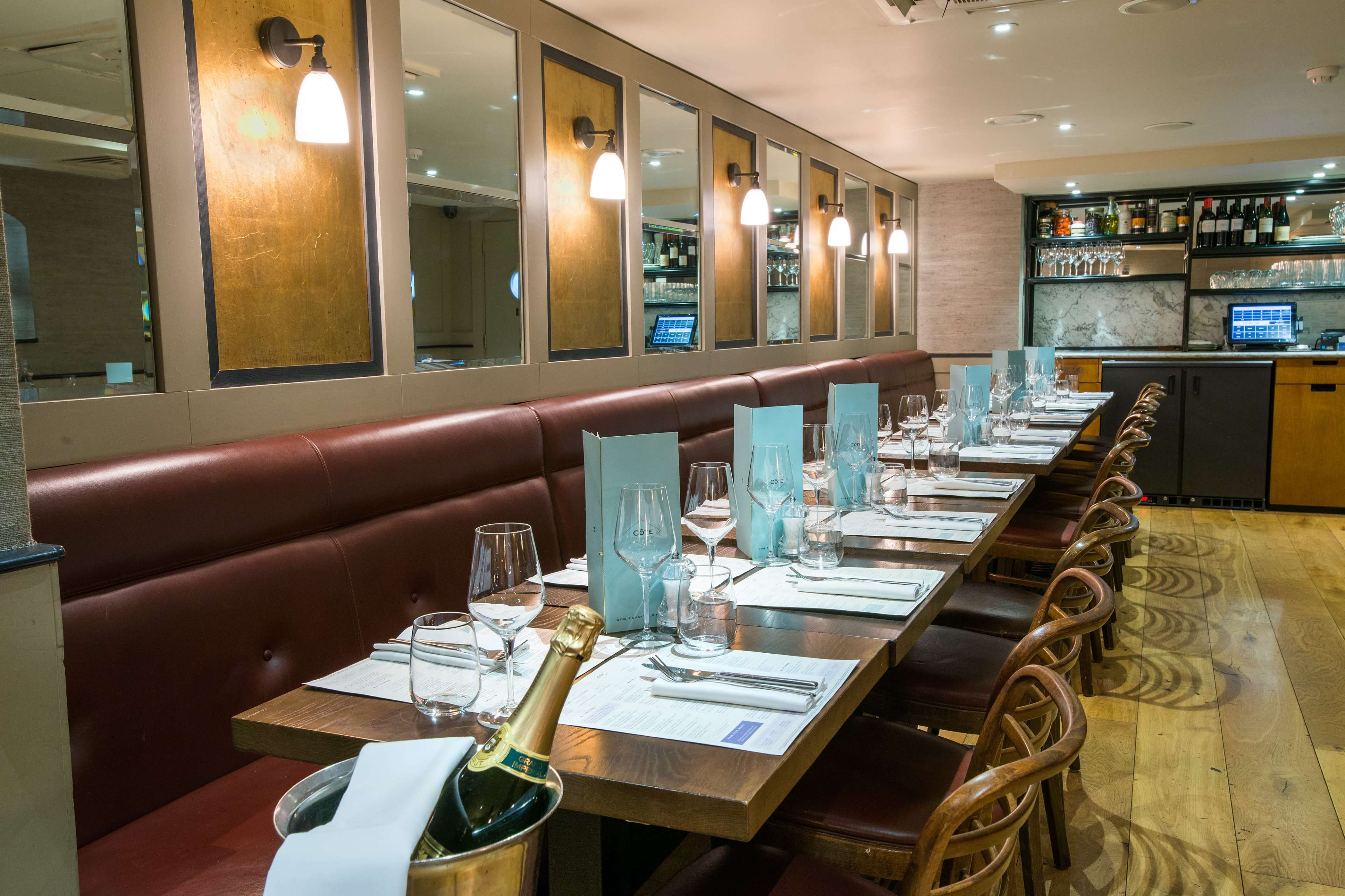 A long wooden dining table is set with glasses, silverware, and menus, surrounded by brown leather benches in a modern restaurant.
