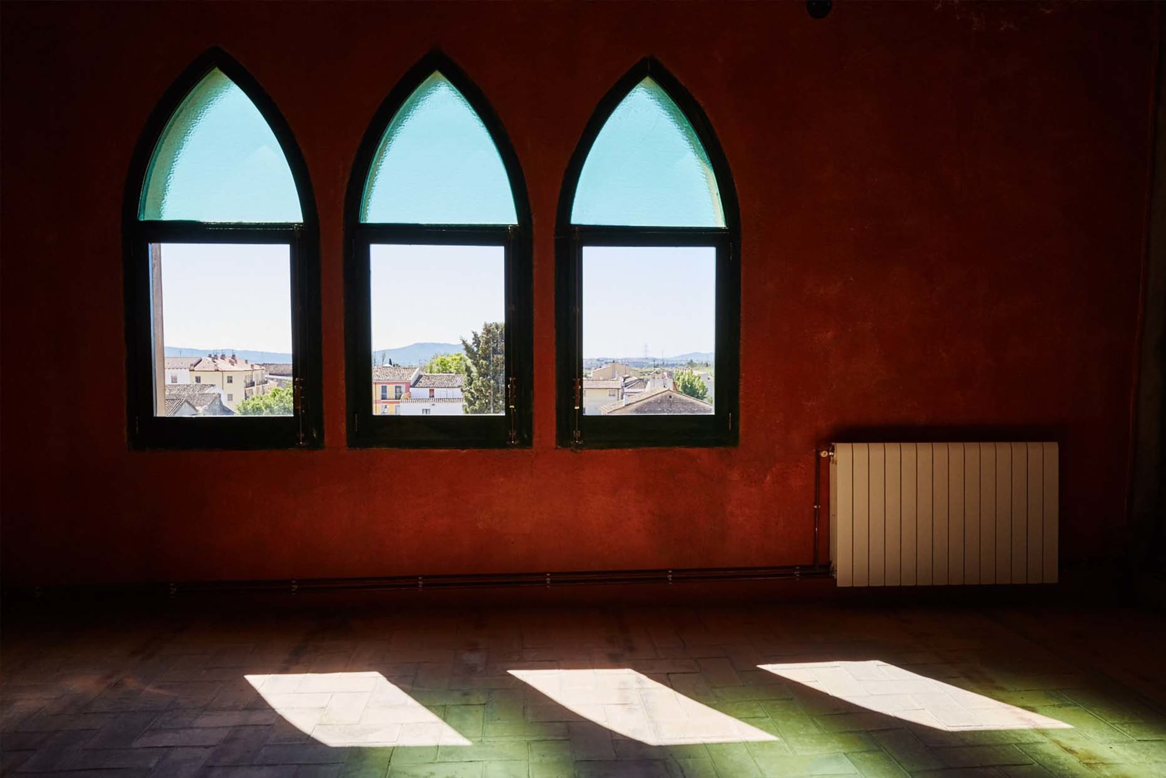 Three arched windows with green frames and glass project sunlight onto a red wall, illuminating the floor in a room overlooking a town.