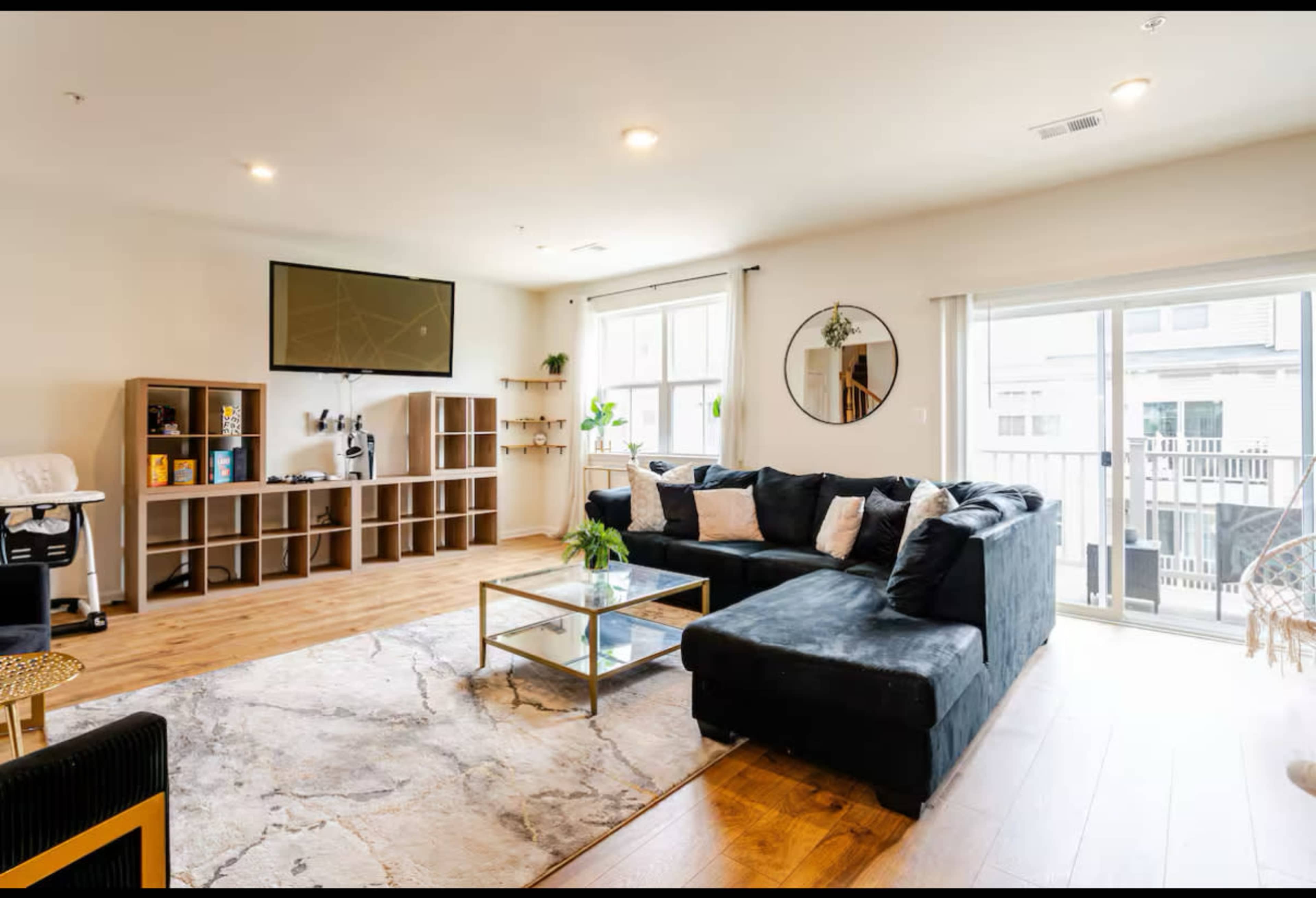 The living room features a sectional sofa, a glass coffee table, a wall-mounted television, and large windows that provide natural light.