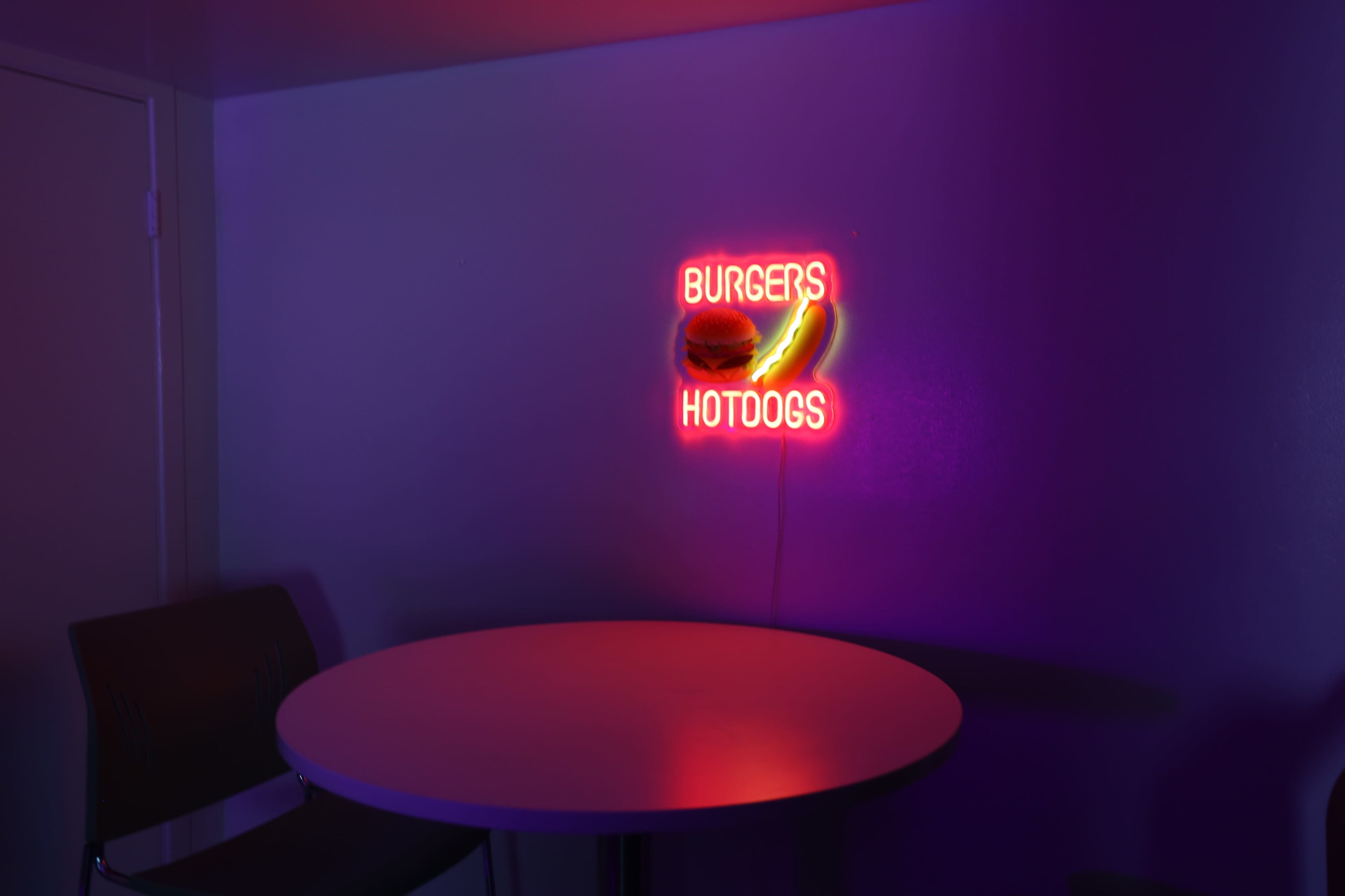 A neon sign displaying "BURGERS" and "HOTDOGS" illuminates a round table in a dimly lit room.