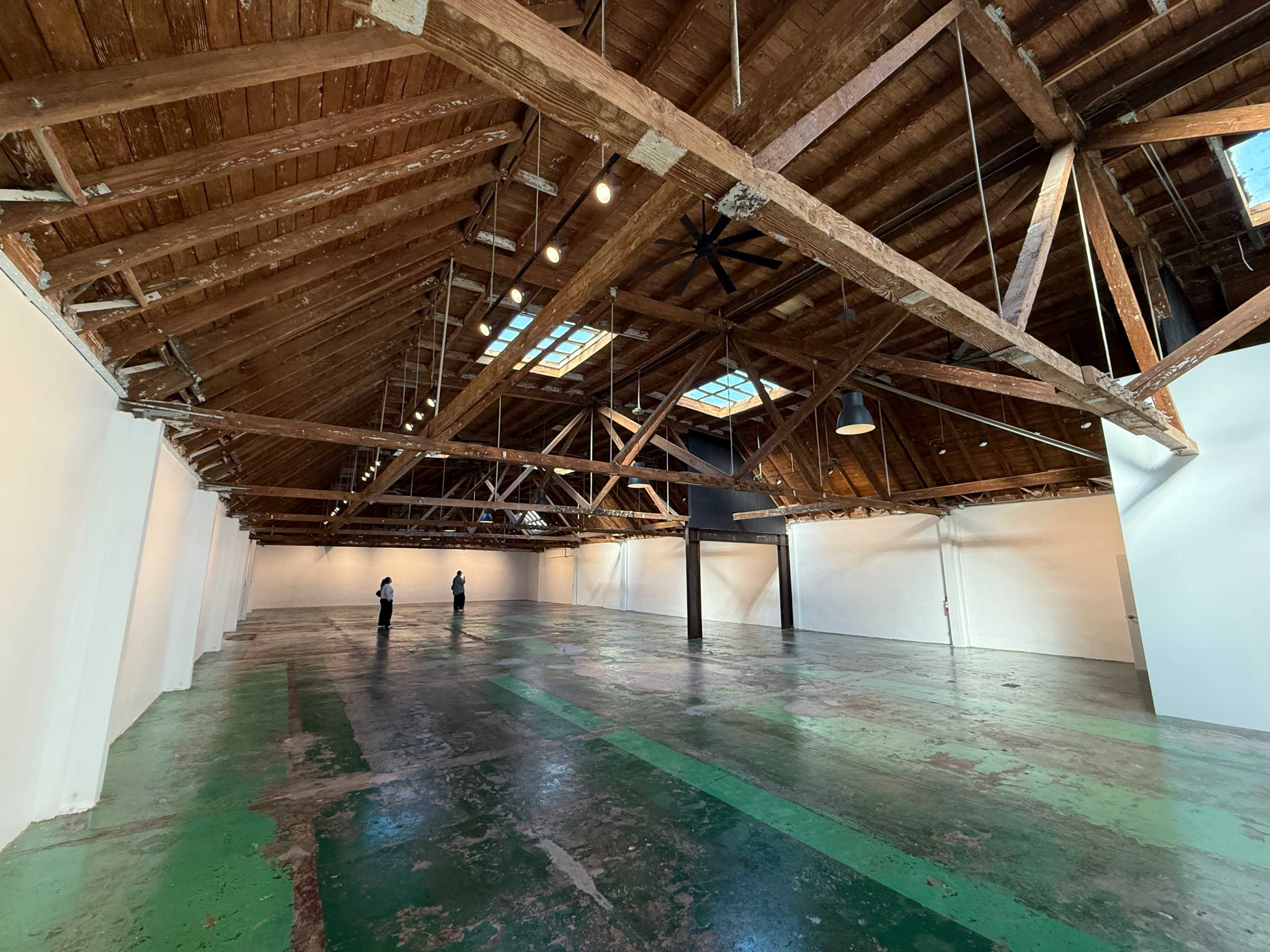 The image shows a spacious warehouse interior with a high, wooden-beamed ceiling and two people standing on a green and gray patterned floor.
