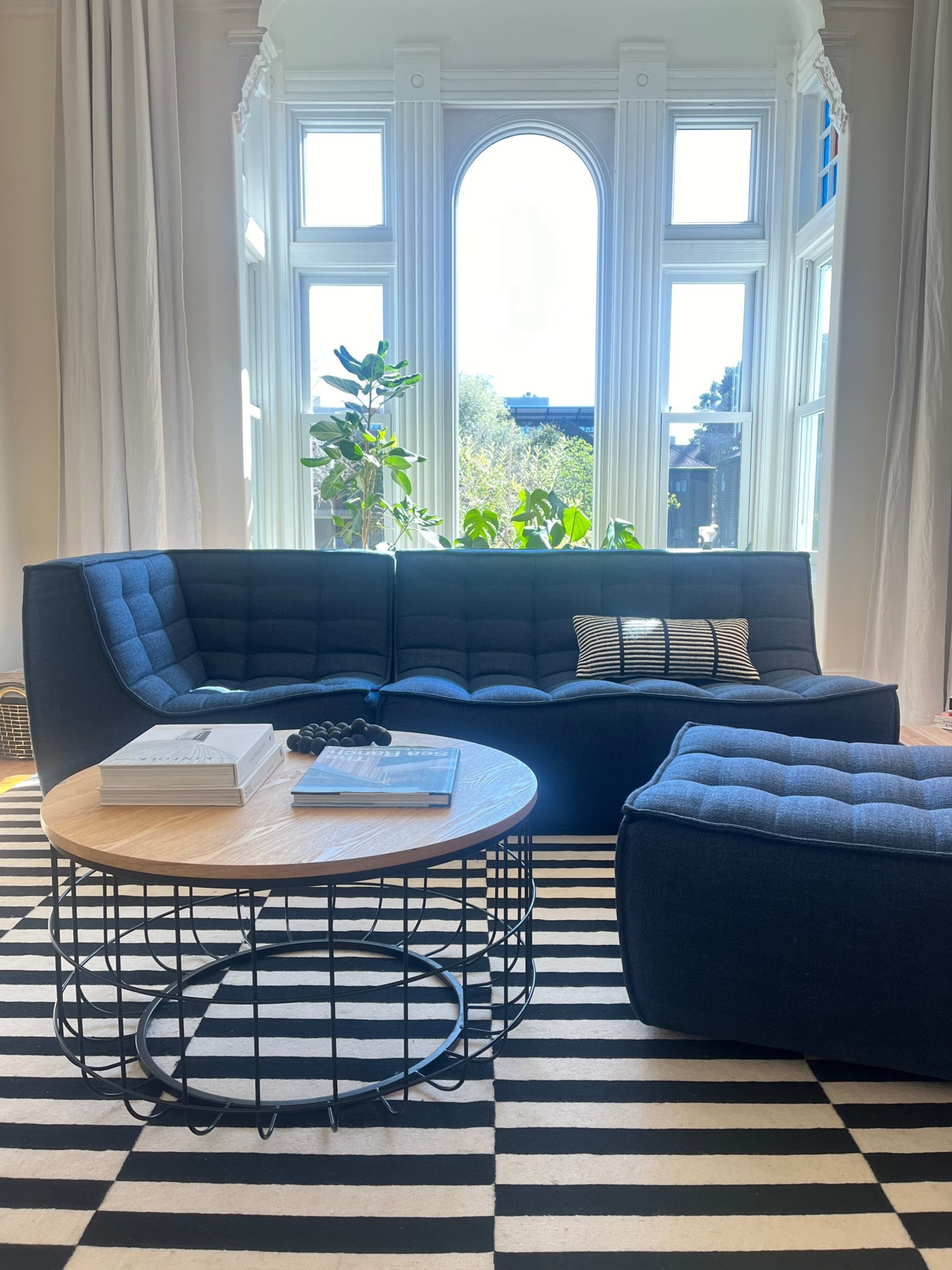 A modern living room features a blue sectional sofa, a round coffee table, and a striped rug, with large windows allowing natural light to enter.