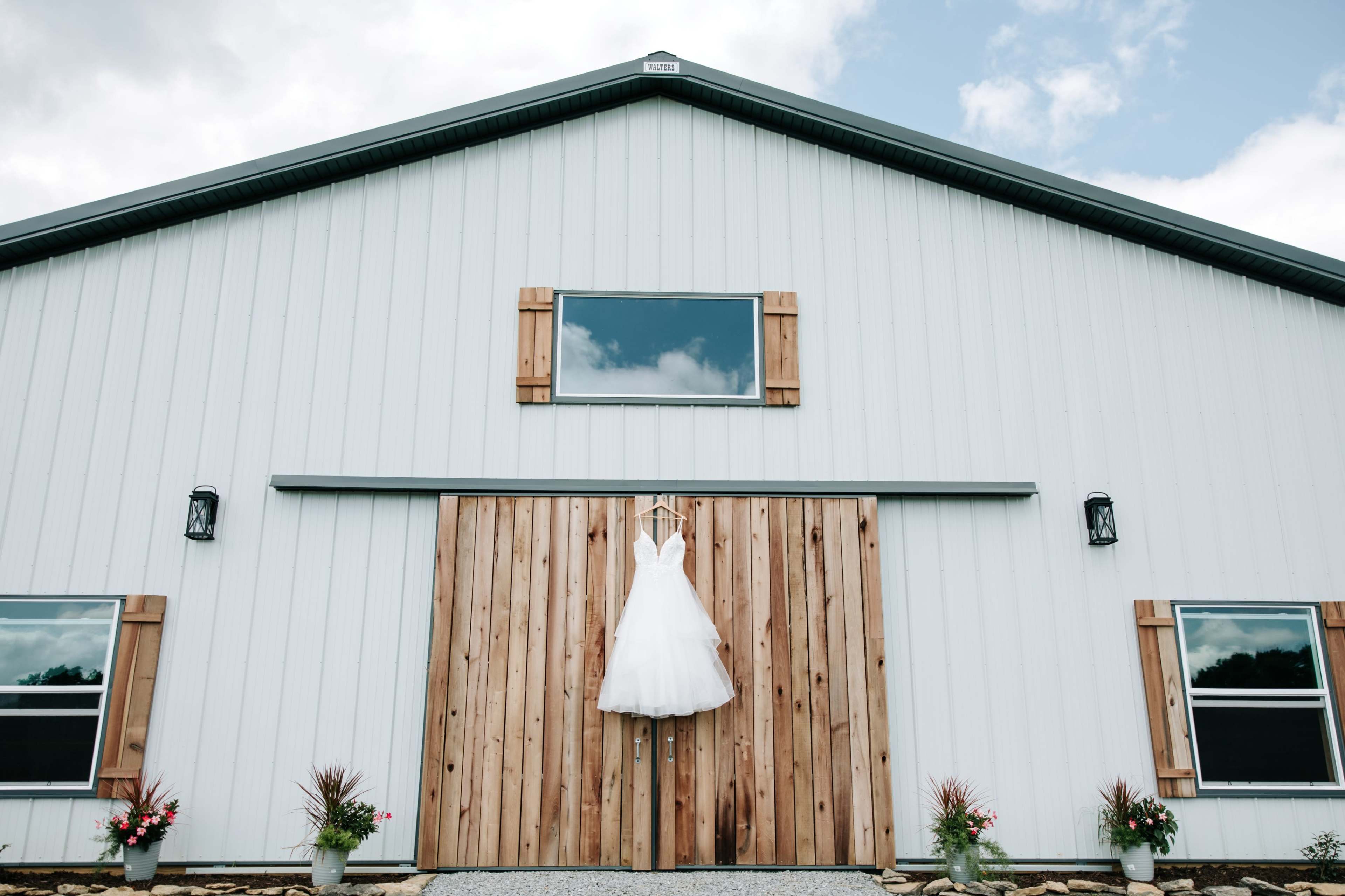 A white wedding dress hangs from a wooden barn door against a gray and white barn facade.