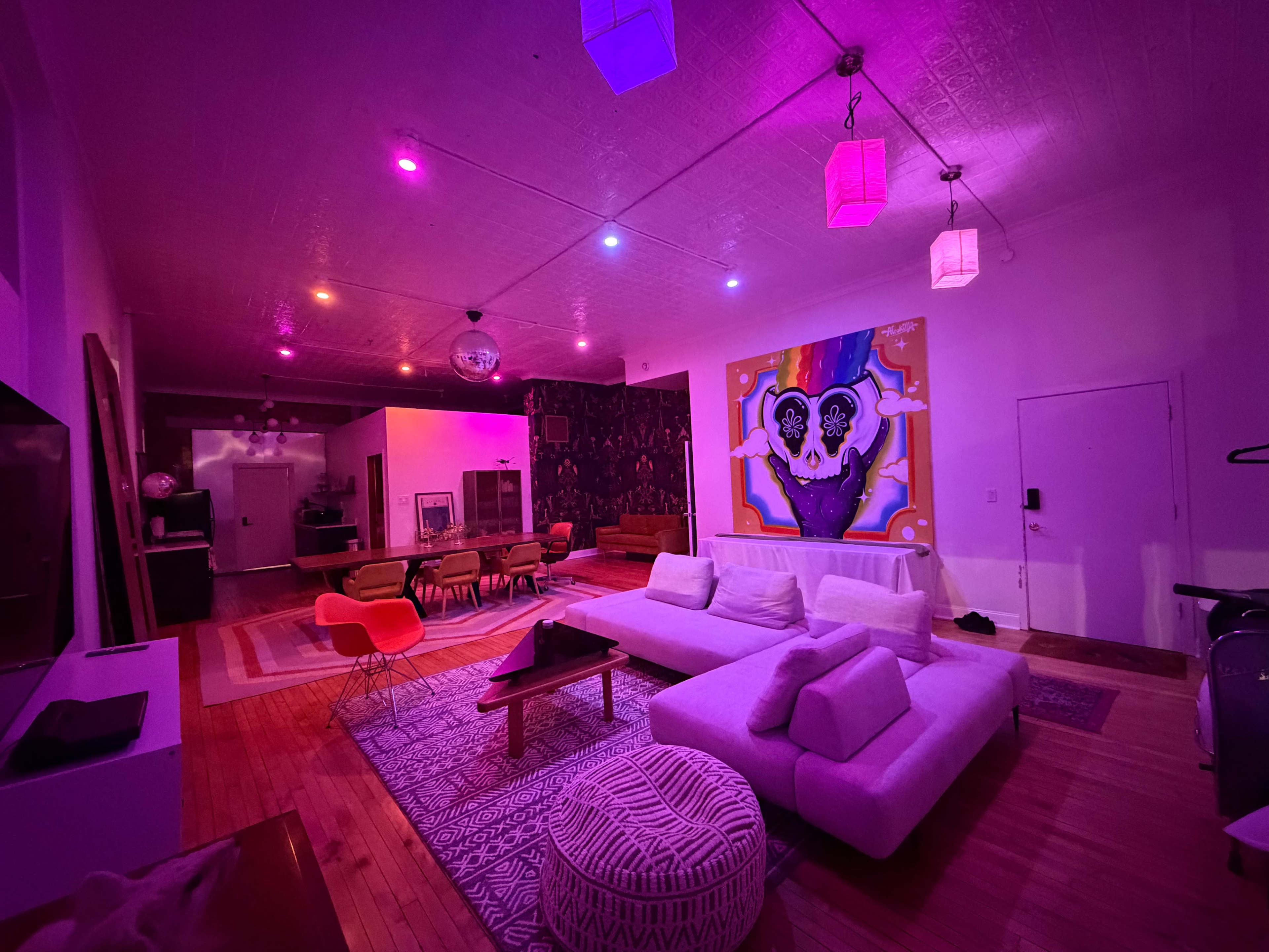 A brightly lit living space features a white sectional sofa, colorful chairs, a large wall artwork, and various lighting fixtures casting a pink hue throughout the room.
