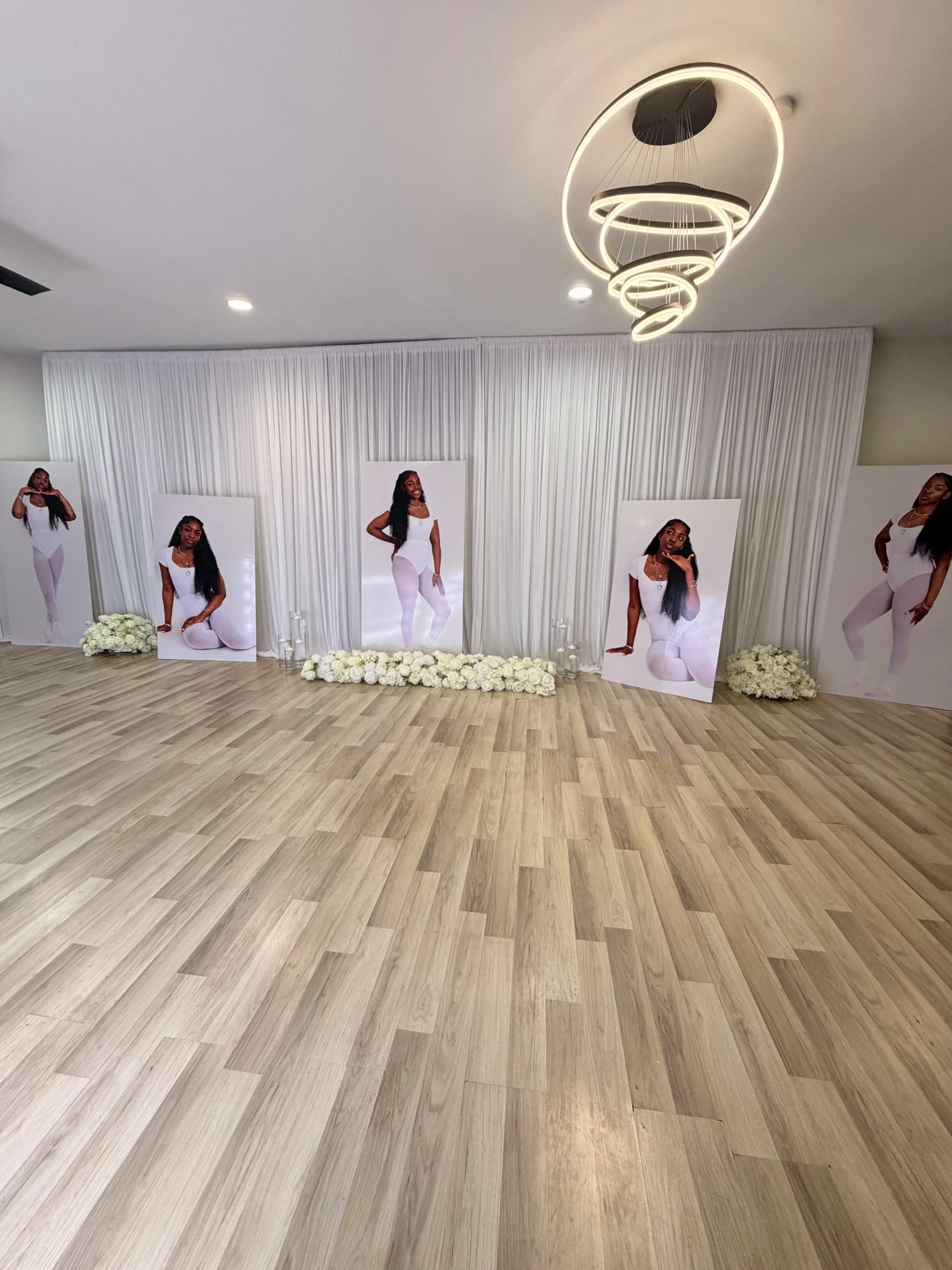 The image shows a spacious indoor setting featuring white draped curtains, four large banners of a woman in white attire, and a floral arrangement on the floor.