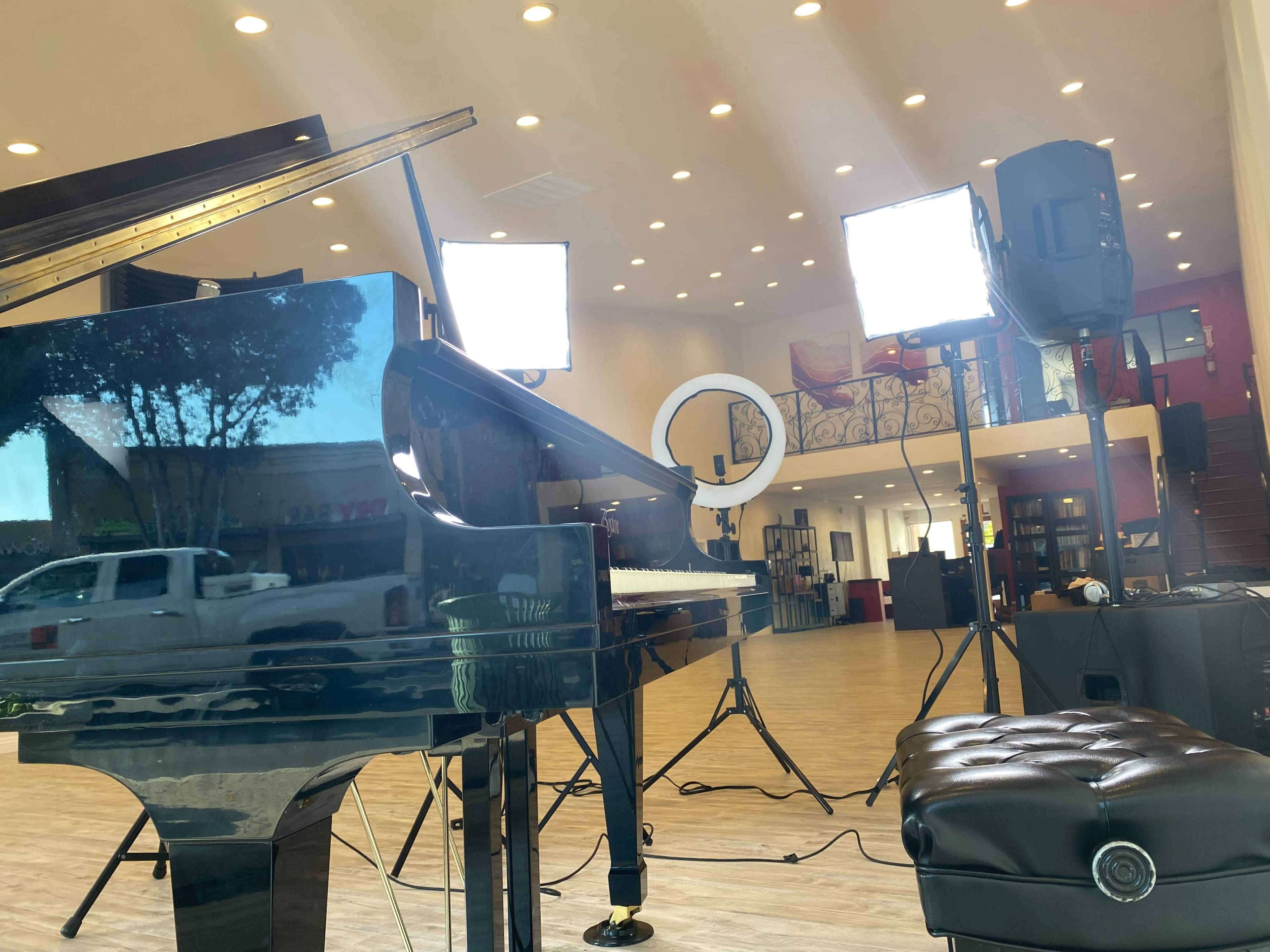 A grand piano is positioned in a well-lit room with studio lights and a camera setup, featuring a view of a parked truck outside.