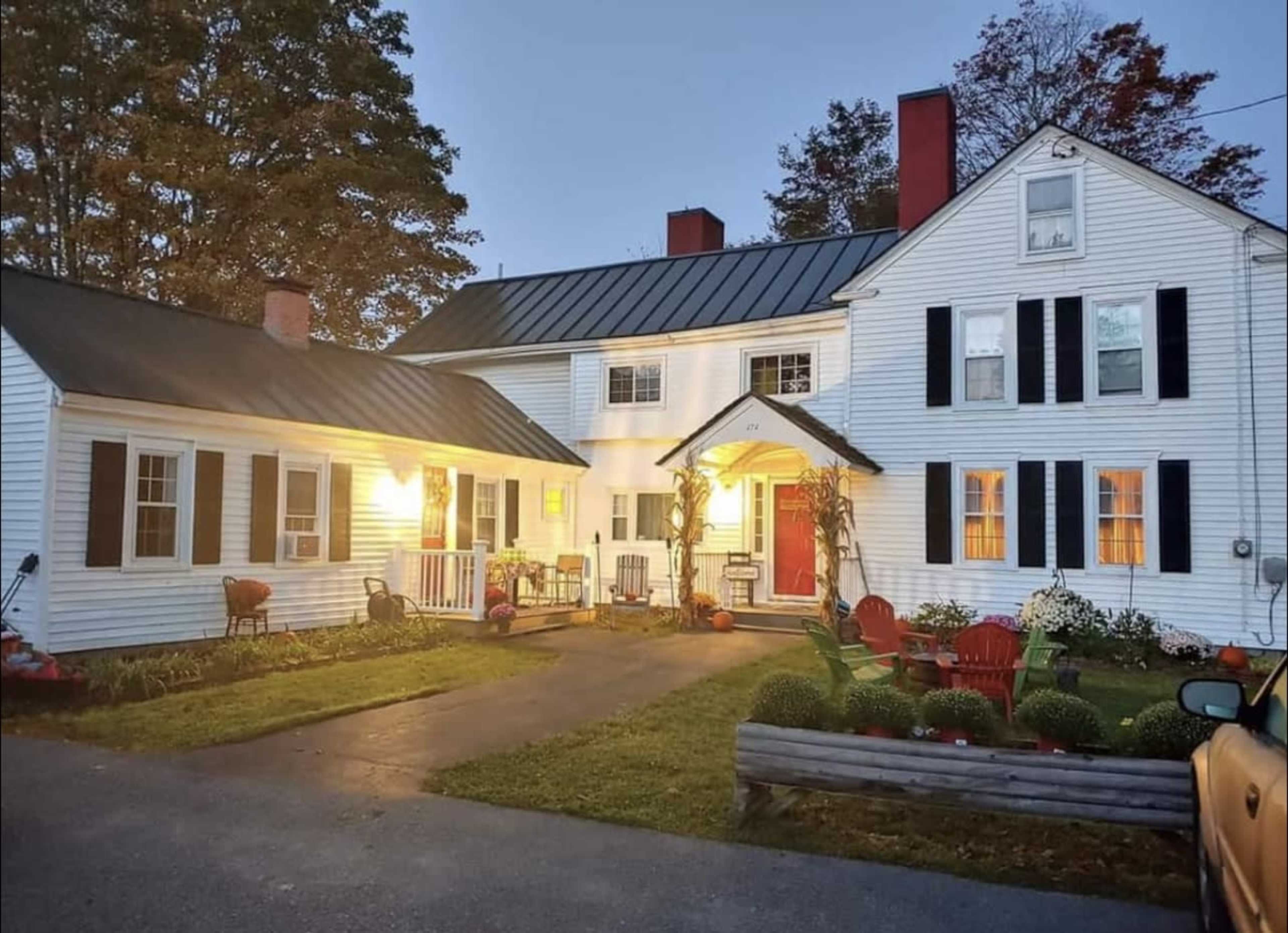 A two-story white house with a black metal roof, a front porch, and a well-maintained yard, illuminated by warm lighting at dusk.