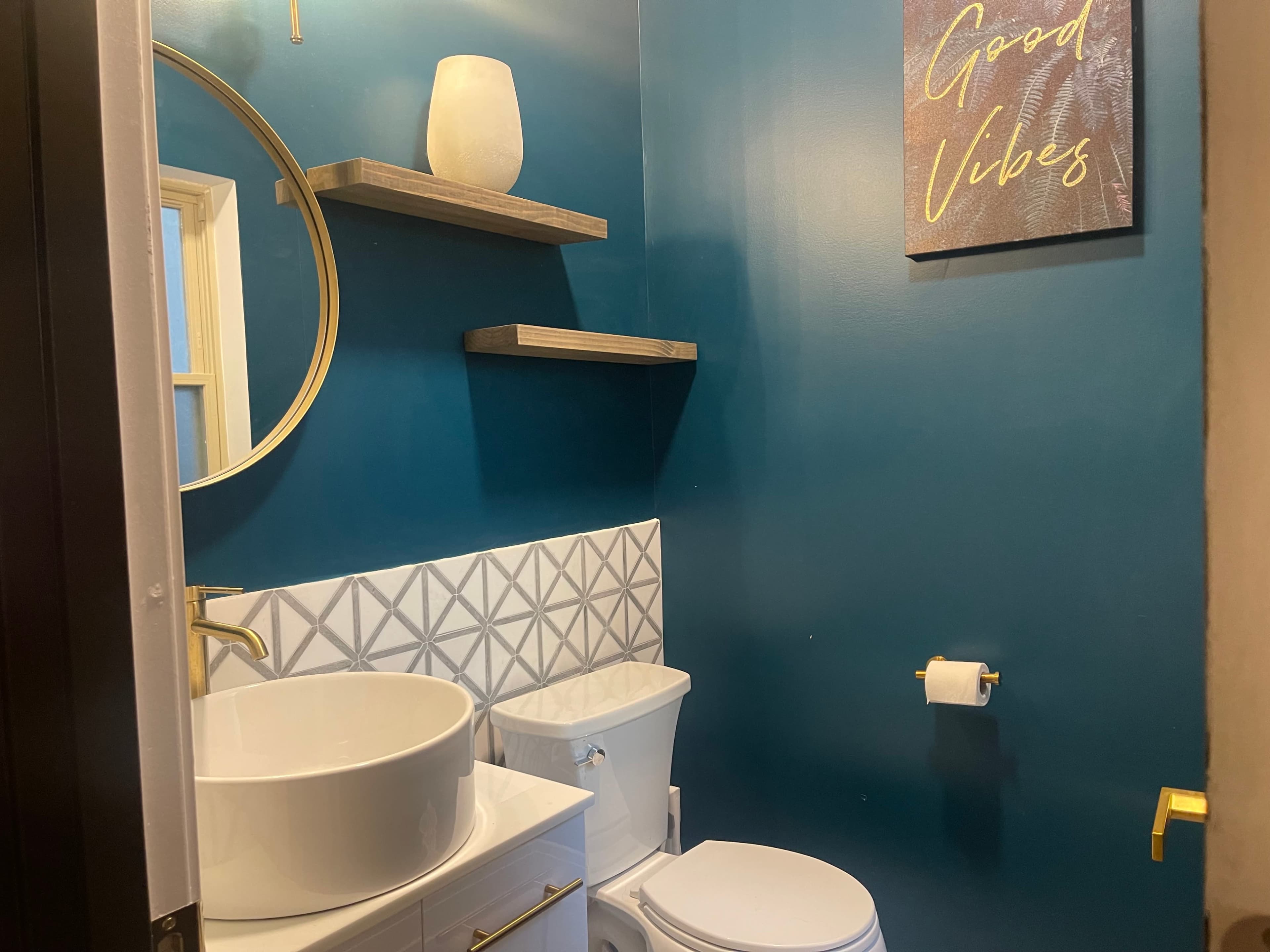 A small bathroom features a round mirror, a white vessel sink on a countertop, a toilet, and teal walls with geometric tiled accents.