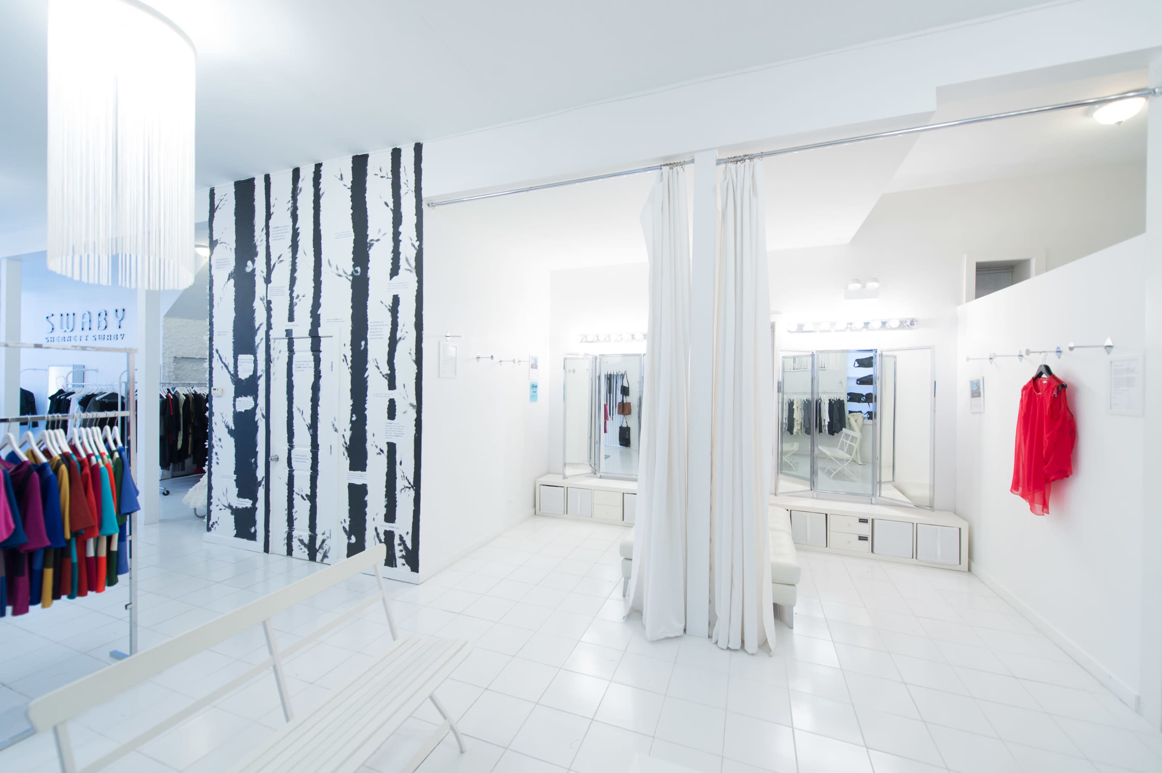 A modern clothing boutique featuring white walls, a patterned accent wall, dressing rooms with curtains, and a seating area with white benches.