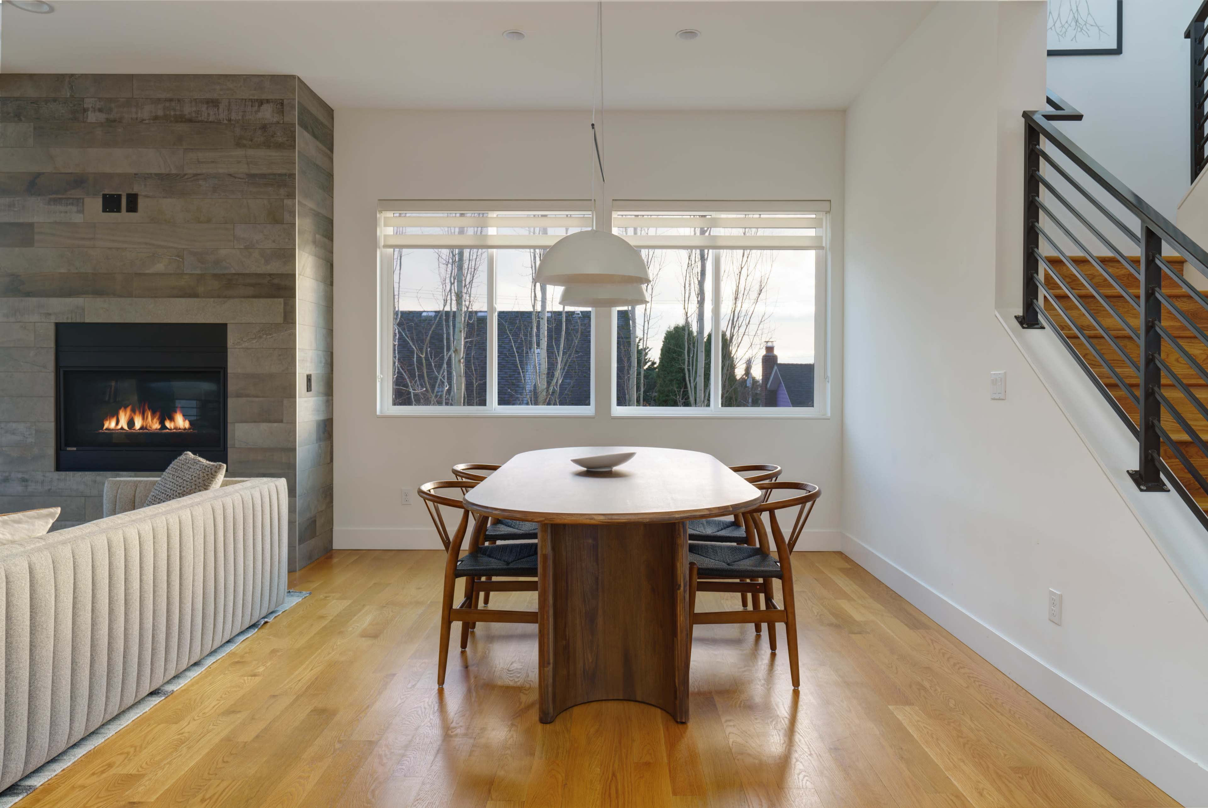 A modern dining area features a round wooden table surrounded by four chairs, with large windows providing natural light and a fireplace nearby.