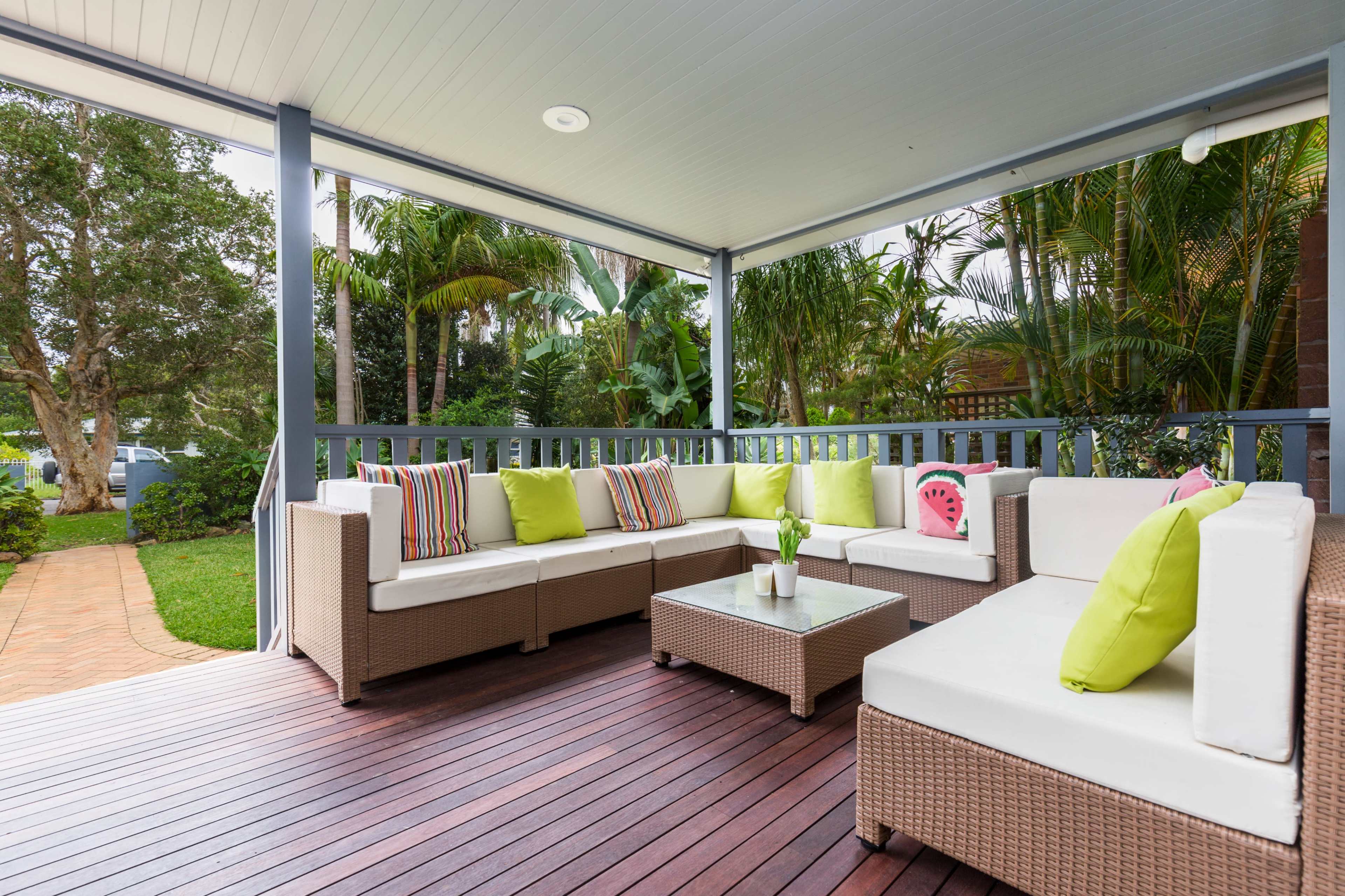 A covered porch features a sectional sofa with brightly colored cushions and a coffee table surrounded by greenery.