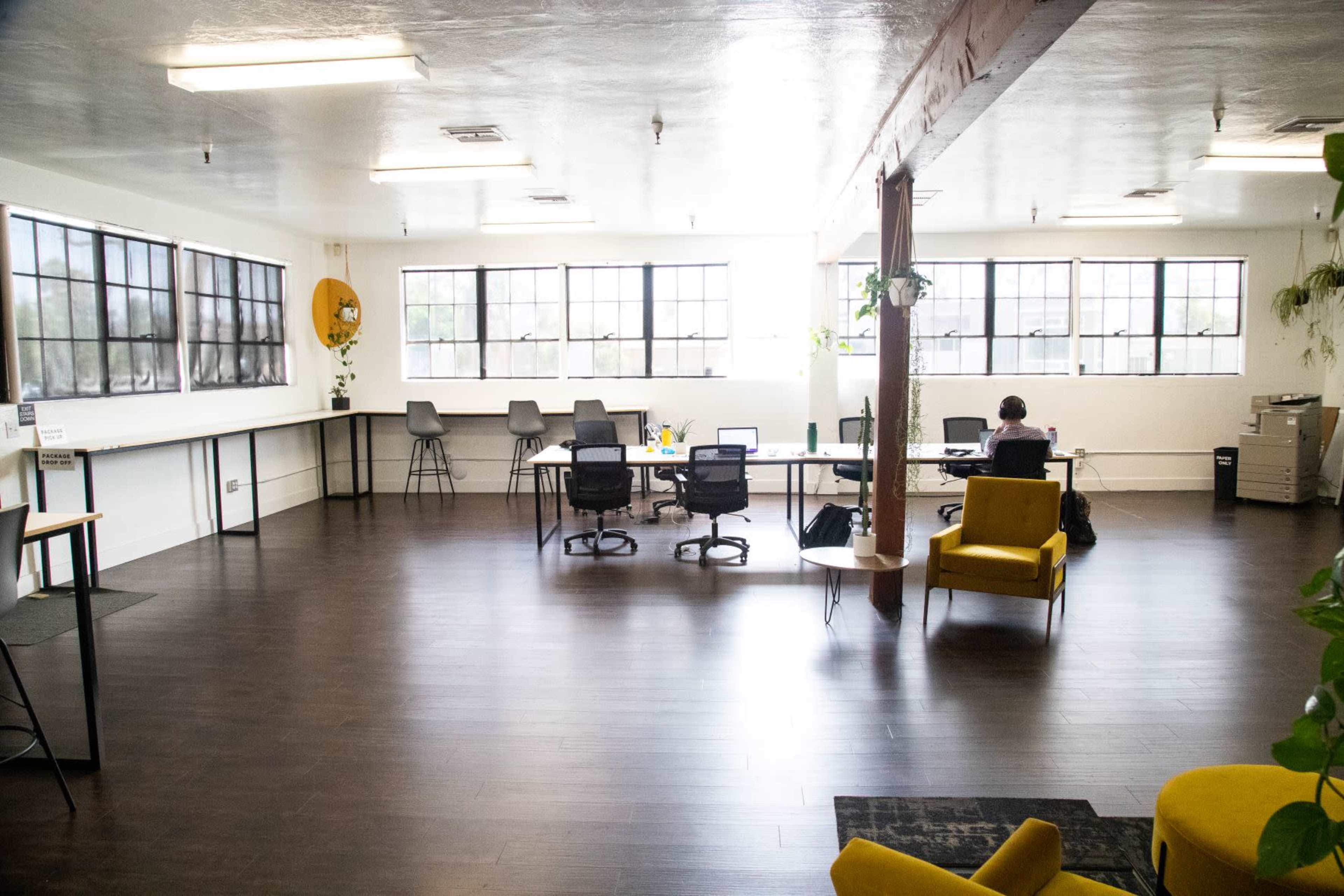 The image shows a spacious office interior with desks, chairs, and large windows allowing natural light.