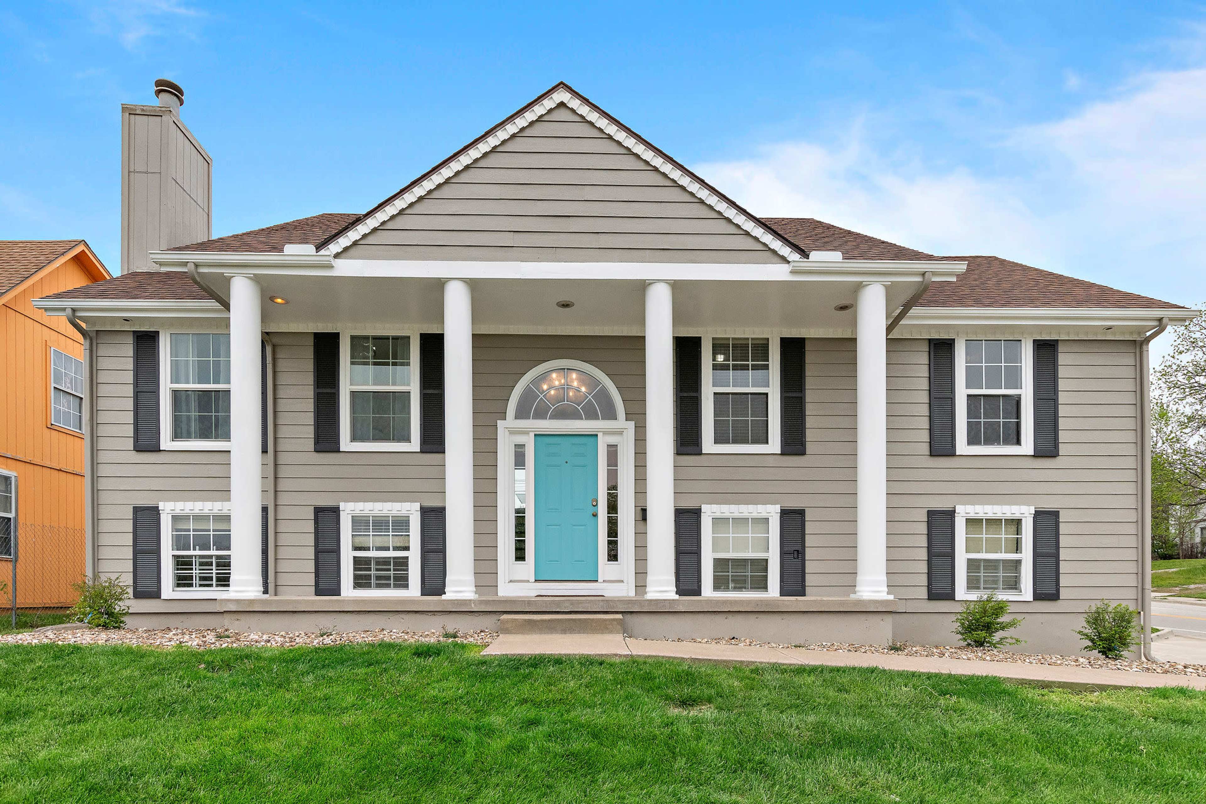 A two-story gray house with white columns and a turquoise front door, surrounded by a well-maintained lawn.