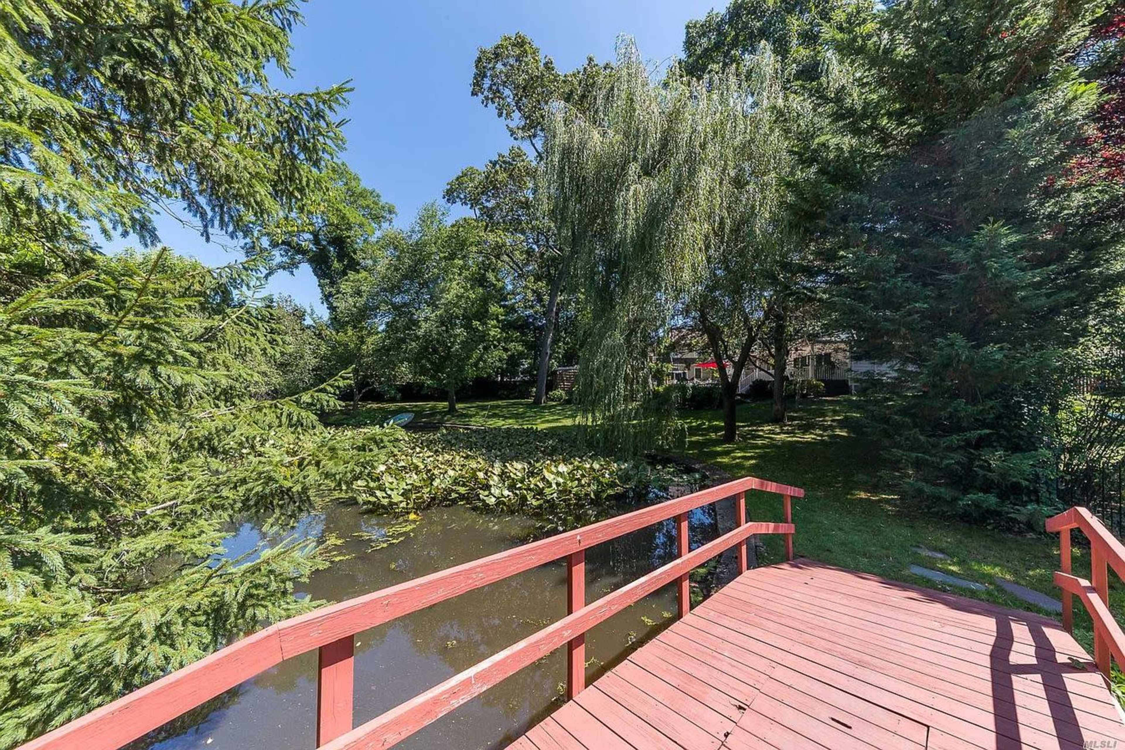 A wooden bridge spans a small pond, surrounded by lush greenery and trees in a tranquil setting.